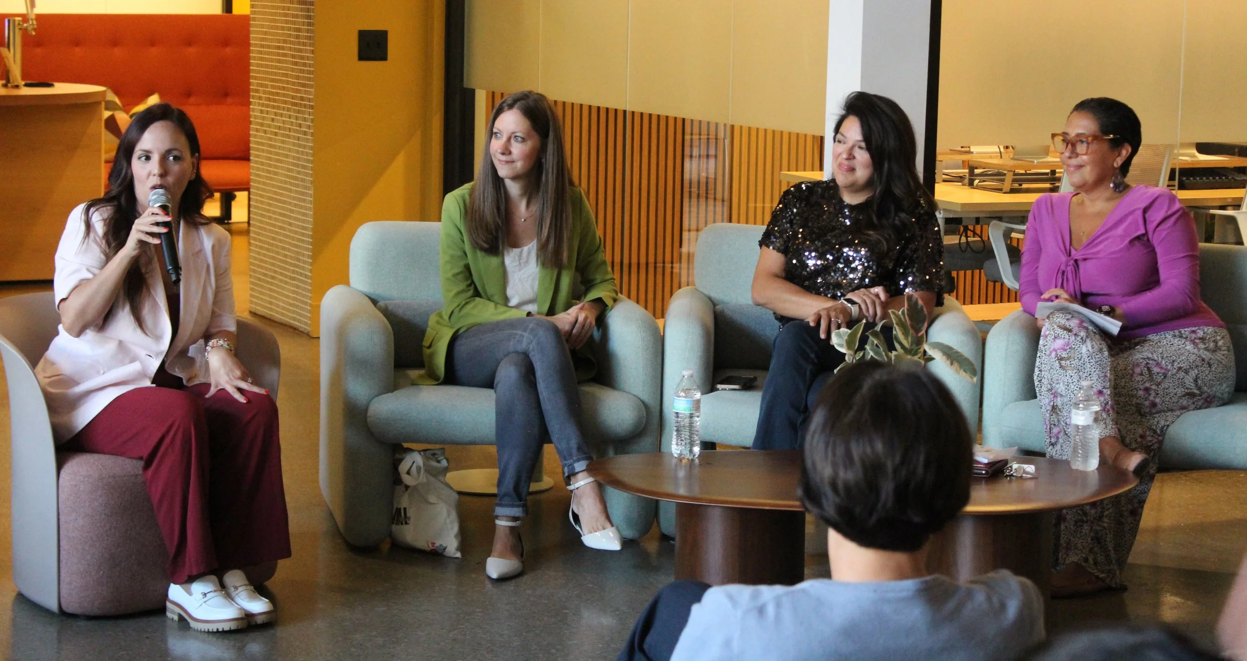 Four women sitting in a semi-circle during an indoor panel discussion or interview. One woman is speaking into a microphone, while the others listen. Water bottles and personal items are on the table in front of them.