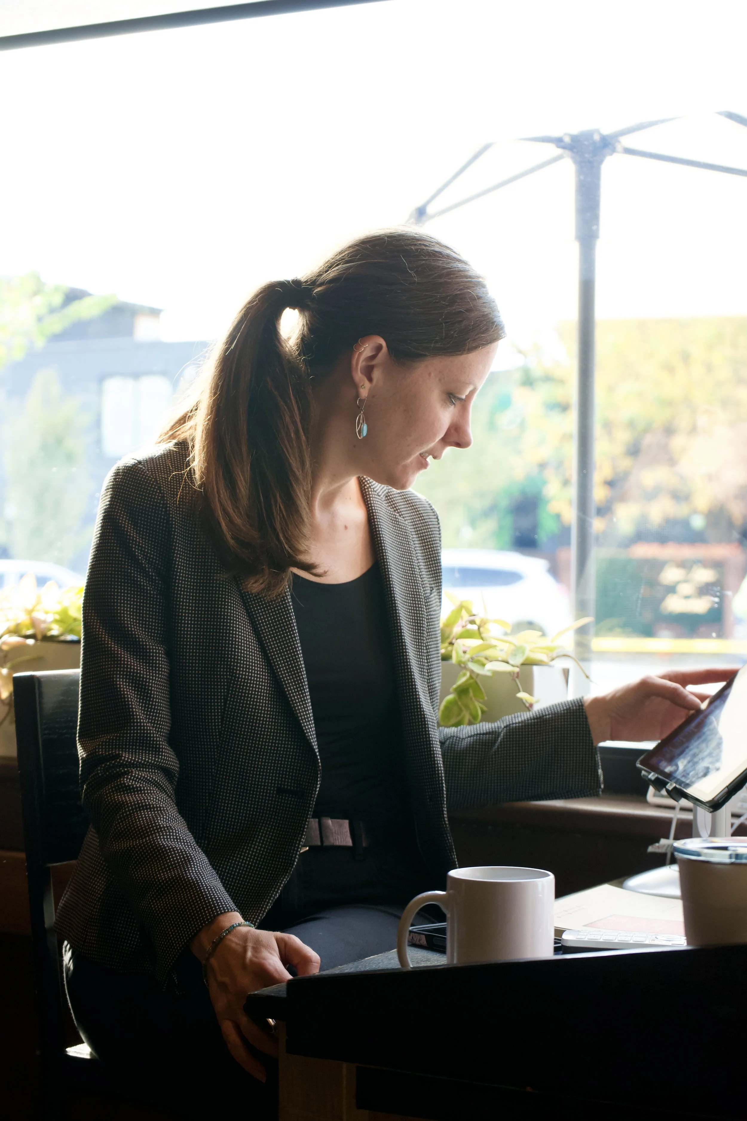 A woman with brown hair tied in a ponytail wearing earrings and a checked blazer sitting at a table with a white mug, a tablet, and a keyboard, looking at a screen.
