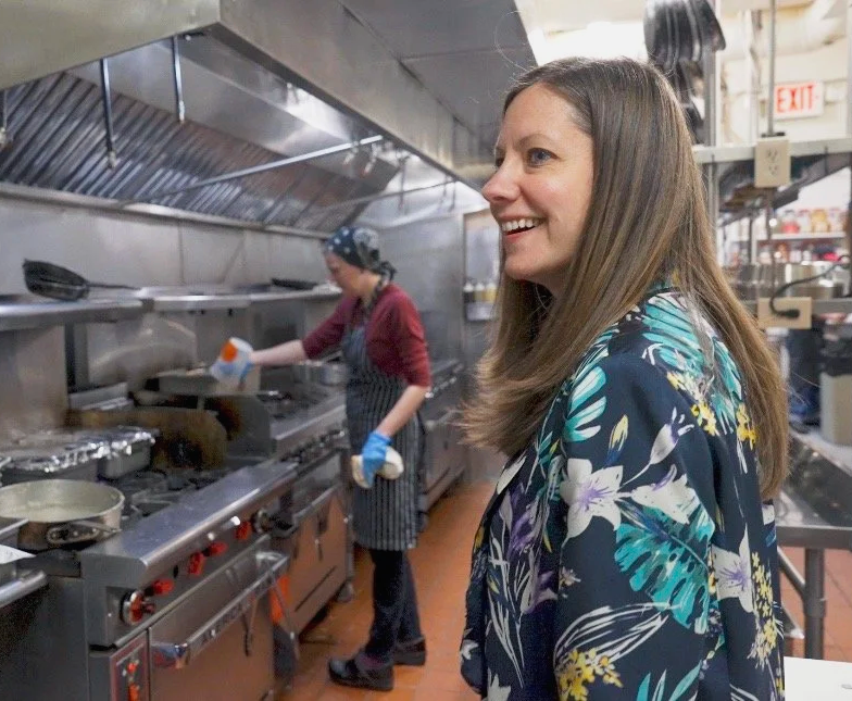 A woman in a floral shirt smiling while working in a commercial kitchen with a chef cooking in the background.