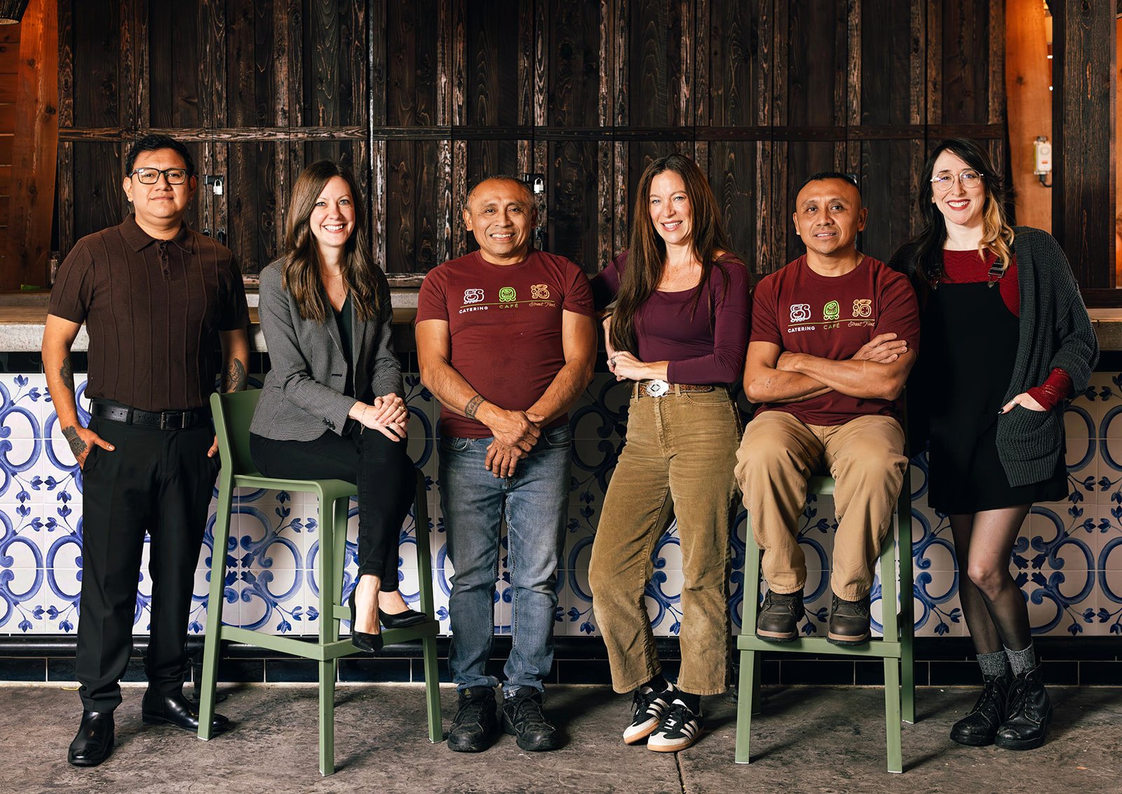 Group of seven diverse people standing and sitting in a row inside a cafe with dark wood and patterned tiles.