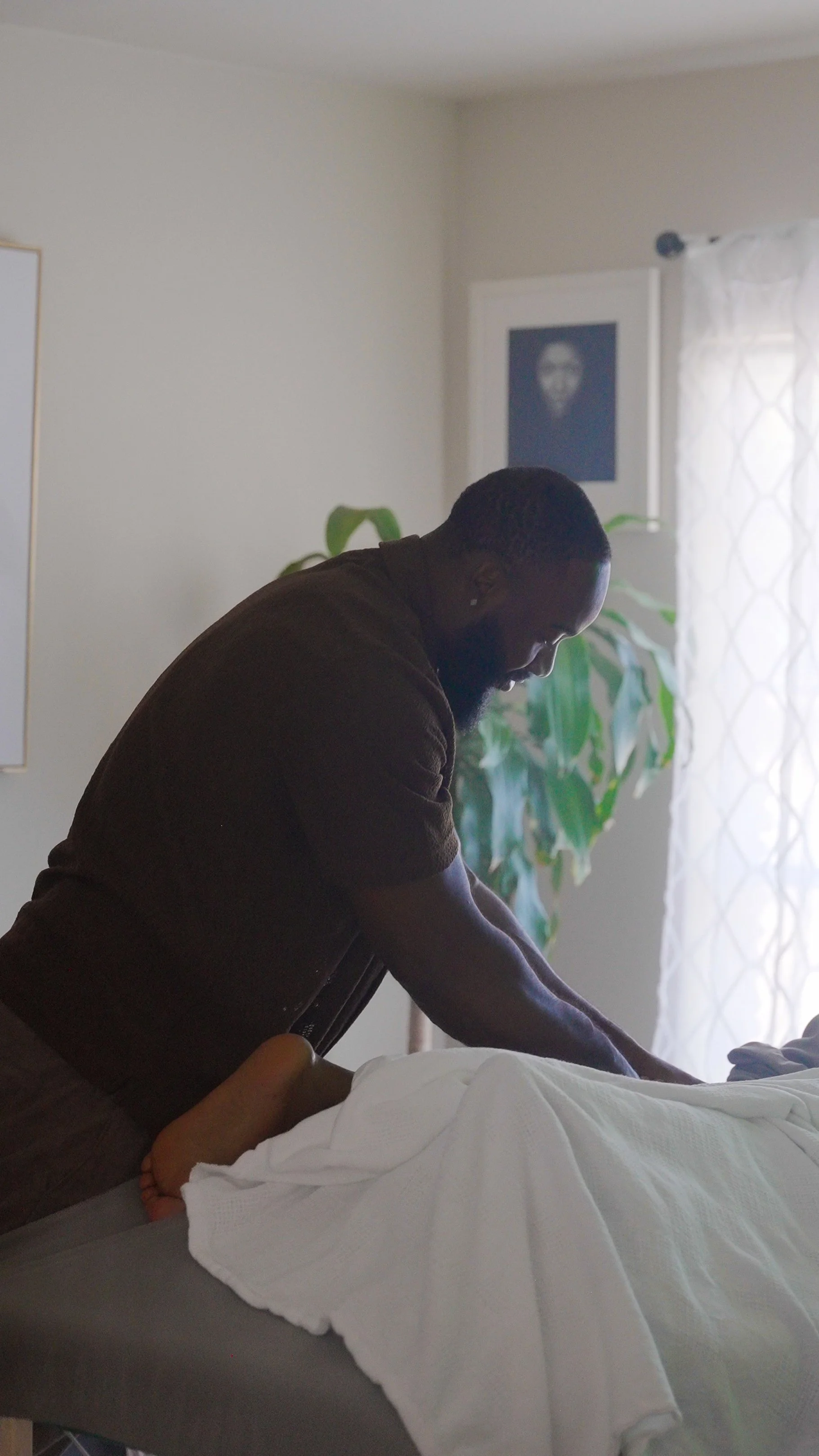A man giving a massage or stretch to another person lying face down on a bed or massage table, in a well-lit room with a plant and a framed picture on the wall in the background. ESO-NETICS massage therapy, holistic bodywork.