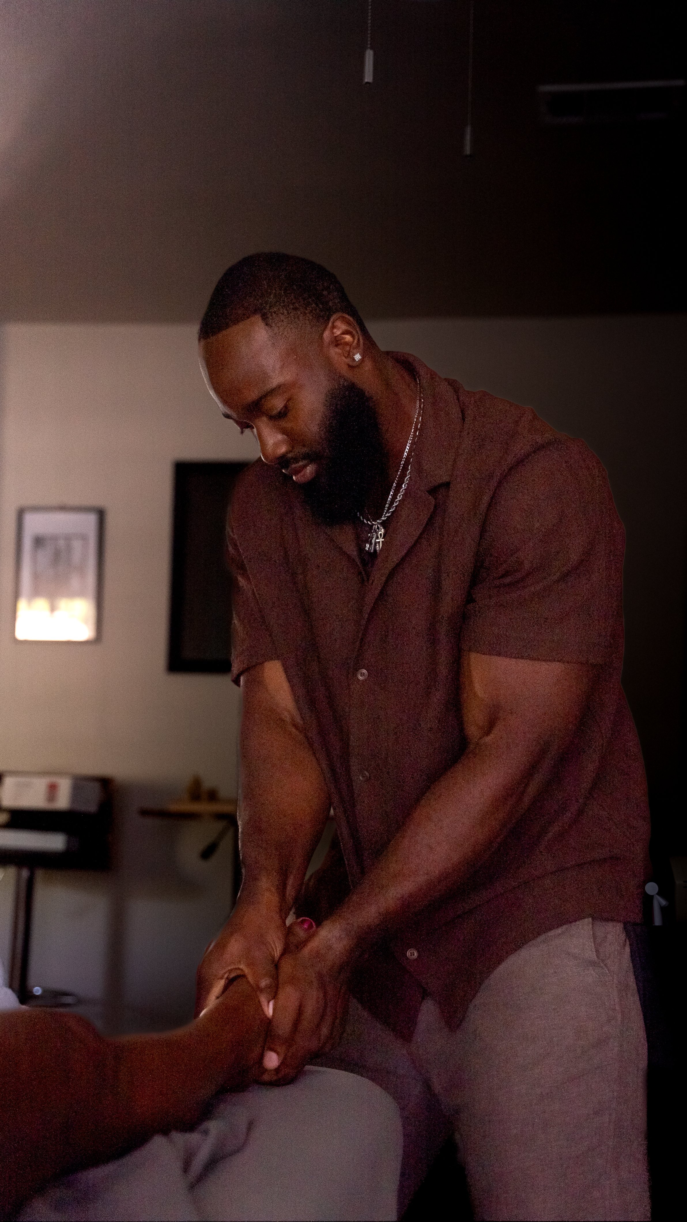 A man with a beard and pierced ear receiving a massage, with focus on his back and head in a dimly lit room.