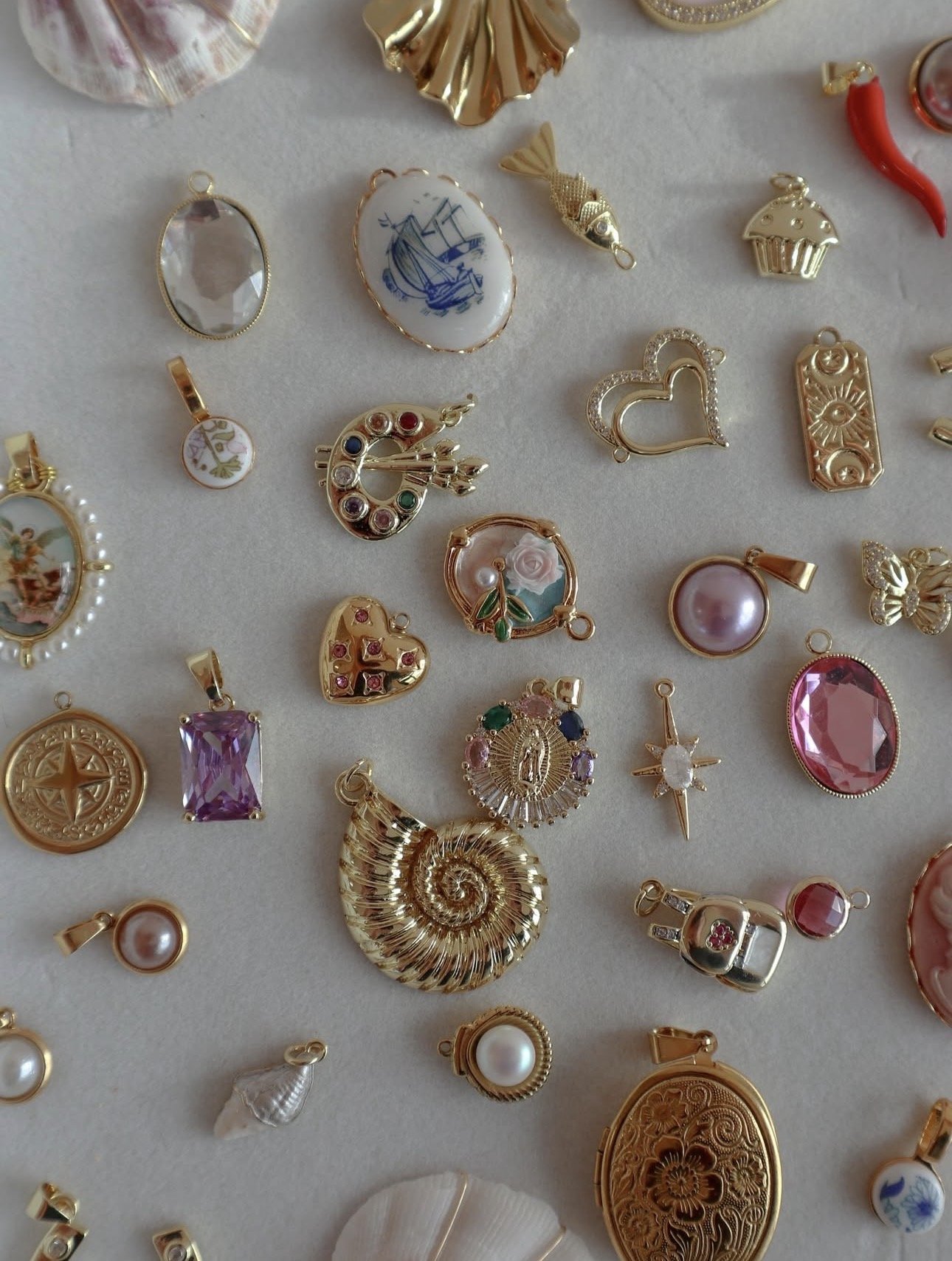 A collection of various jewelry charms and pendants, including shells, hearts, pearls, a compass, a purple gemstone, a spiral shell, a rainbow palette, and other decorative pieces in gold, silver, and colorful designs.