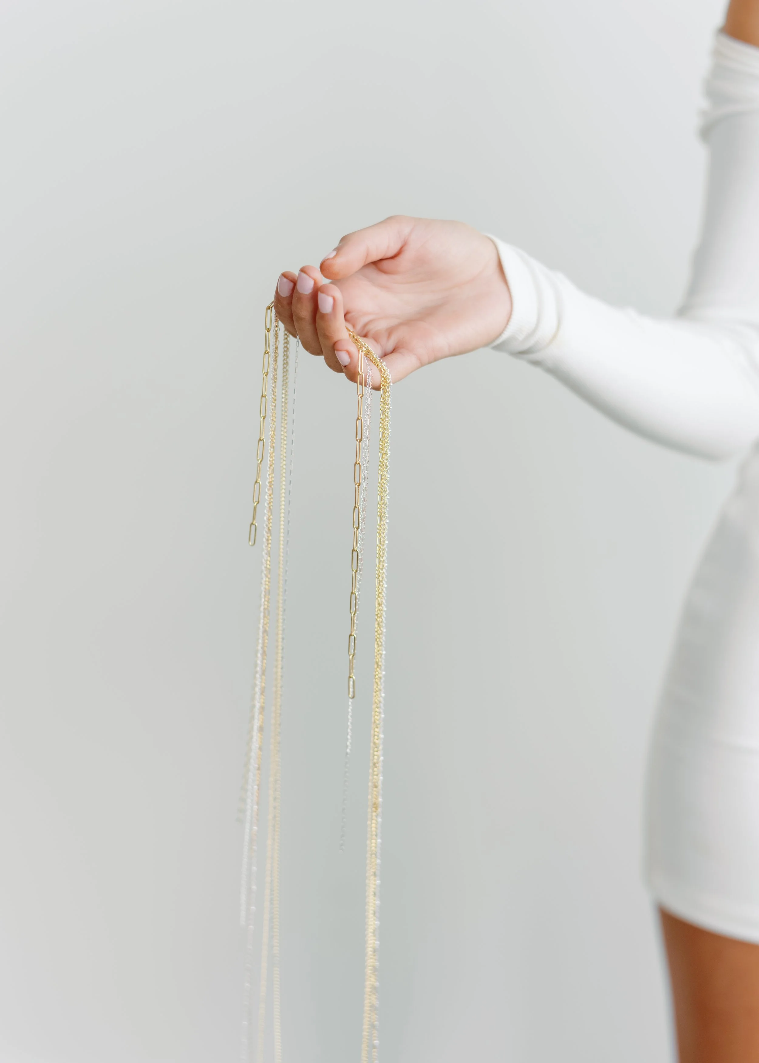 A woman in a white long-sleeve shirt holds several gold and silver chains against a plain light background.