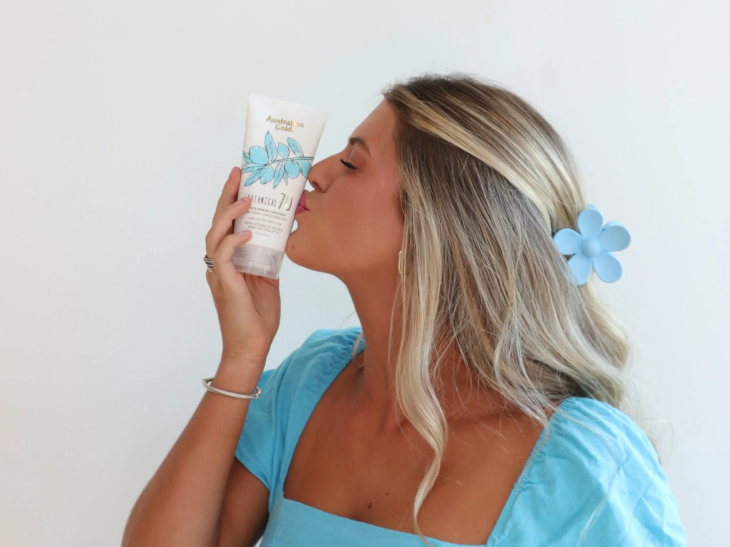 A woman with blonde hair decorated with a blue flower holds a bottle of Australian Gold SPF 70 sunscreen close to her face as if smelling it, wearing a blue top and a silver bracelet.