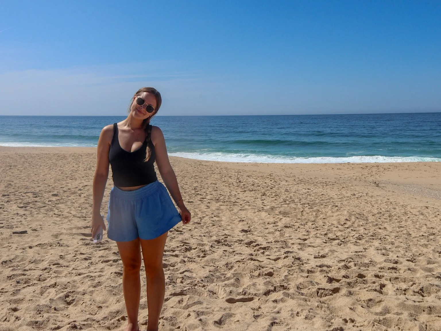 A woman in sunglasses wearing a black tank top and blue shorts stands on a sandy beach with the ocean and blue sky in the background.