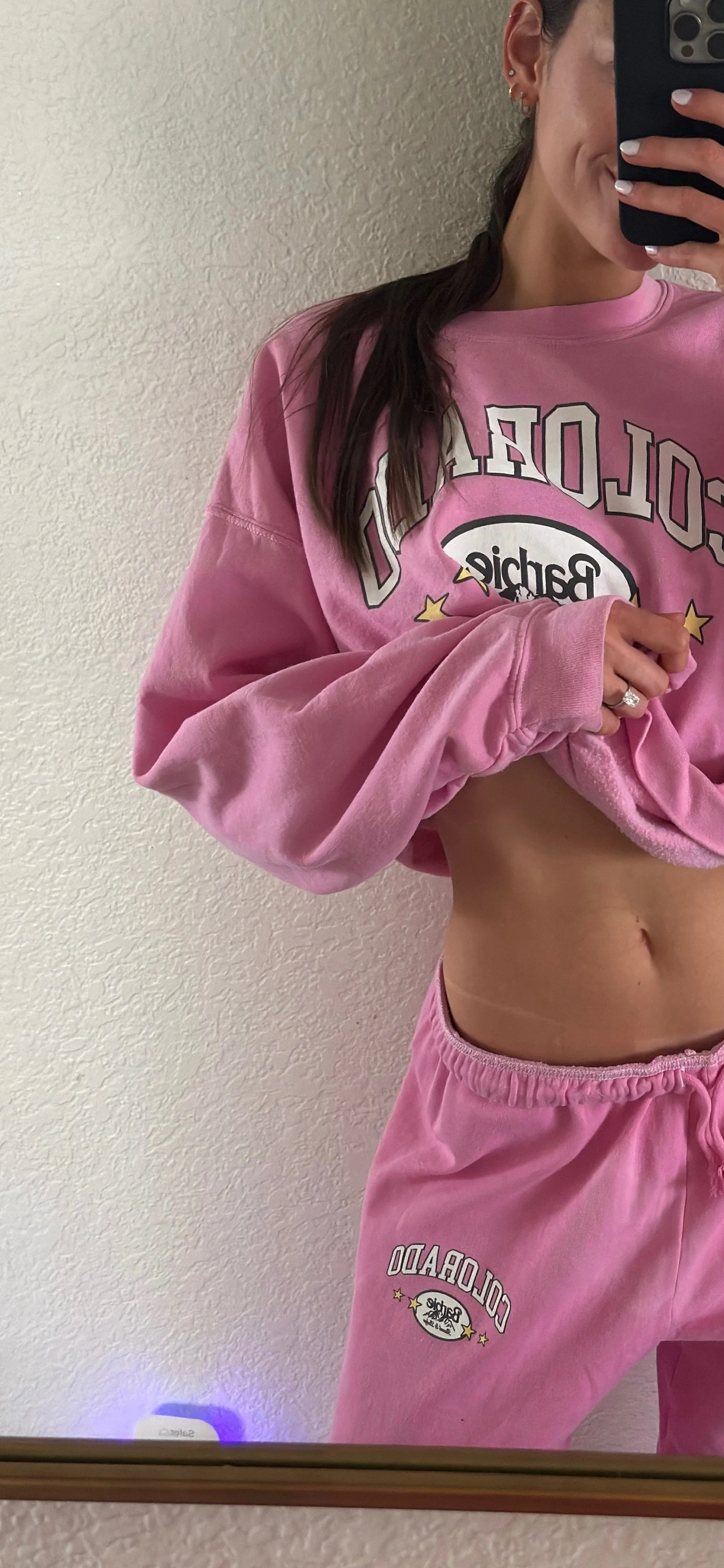 A person taking a mirror selfie wearing a pink sweatshirt and matching pink sweatpants with 'Colorado' and 'Pachie' logos, pulling up the sweatshirt to reveal their midriff.