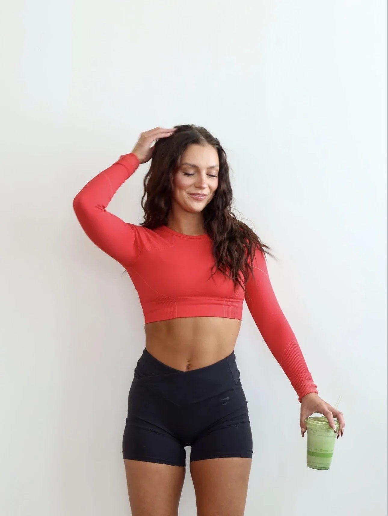 A woman in a red long-sleeve crop top and black shorts holding a green smoothie, standing against a plain white wall.