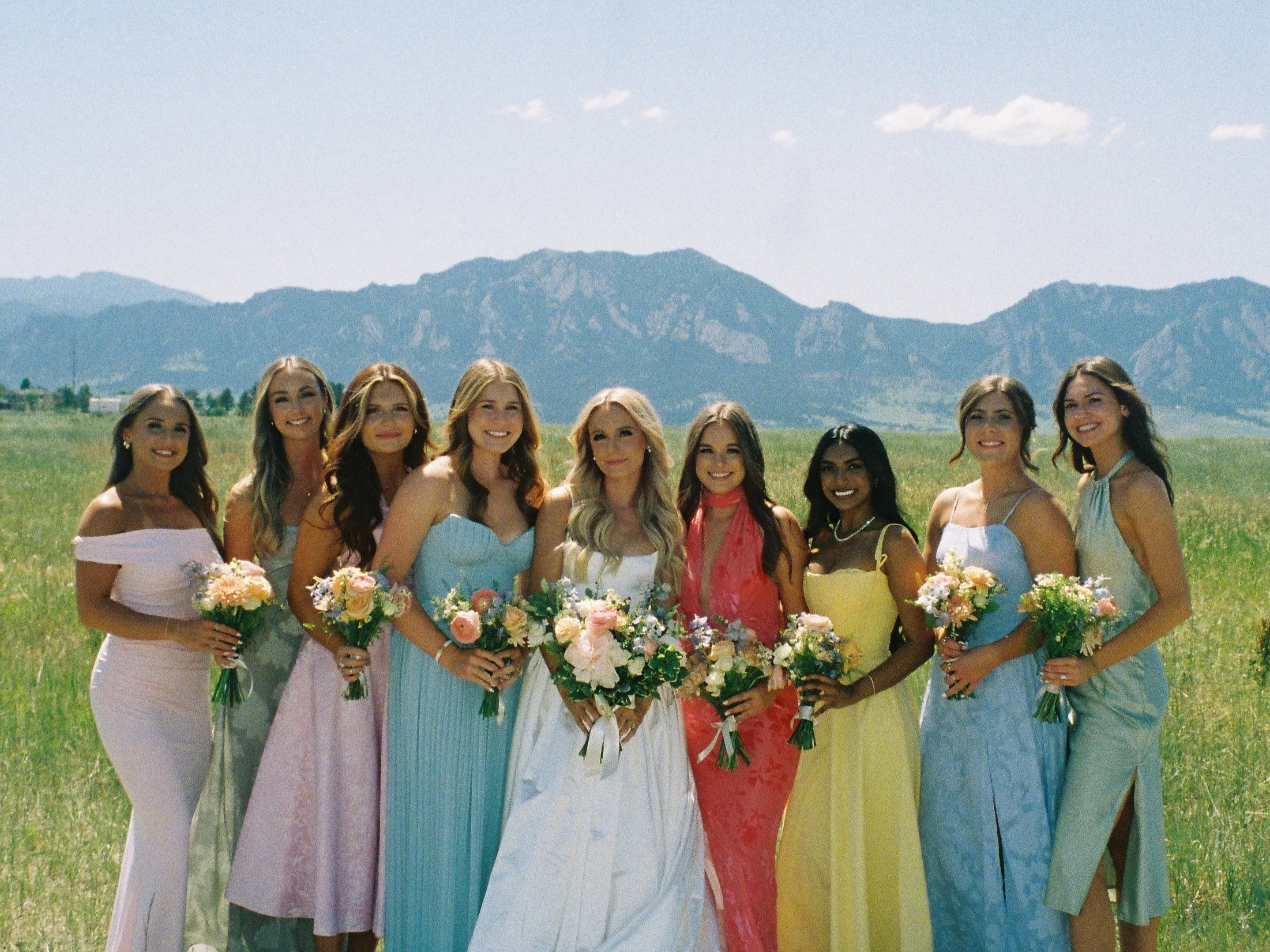 A group of ten young women in colorful dresses, holding bouquets, standing outdoors in a grassy field with mountains in the background, during a sunny day.