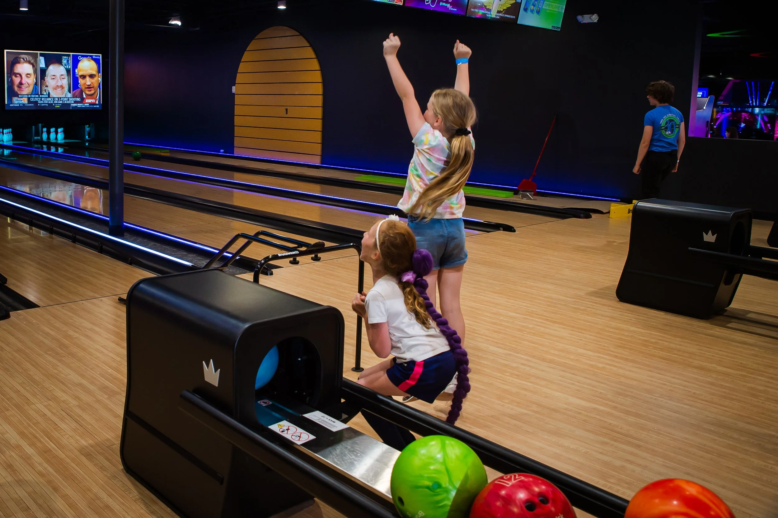 Children at a bowling alley, one girl with arms raised celebrating after a throw, another girl sitting on the floor-looking at the bowling ball, and a boy standing near the lanes. Multiple bowling balls in foreground, television screens displaying sports, dark walls, and wooden lanes in the background.