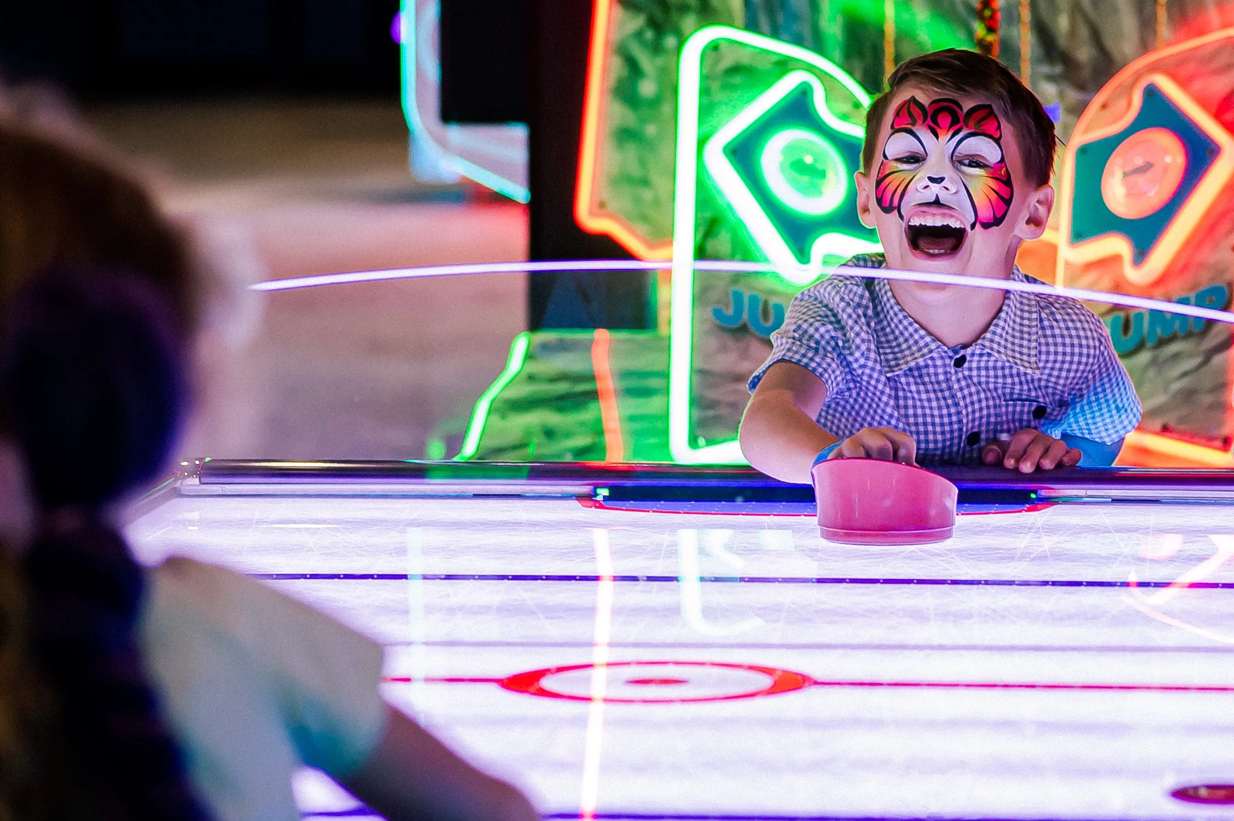 A young boy with face paint resembling a tiger, smiling and leaning on the ice rink barrier at an amusement park or carnival, with bright neon lights in the background.