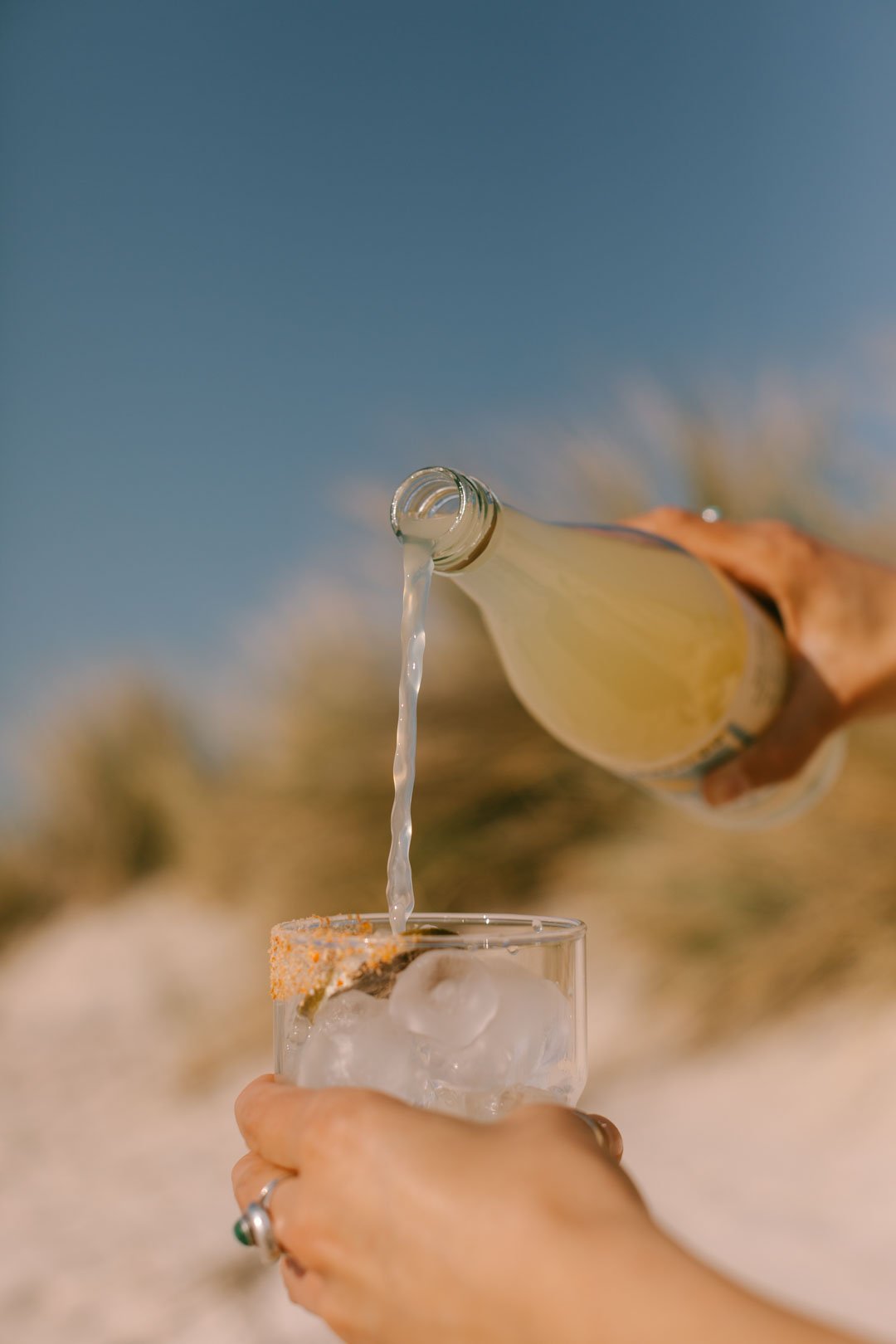 Someone pouring a Salty Fridays Margarita mix from a bottle into a glass with ice, outdoors with a blurred background of trees and blue sky.