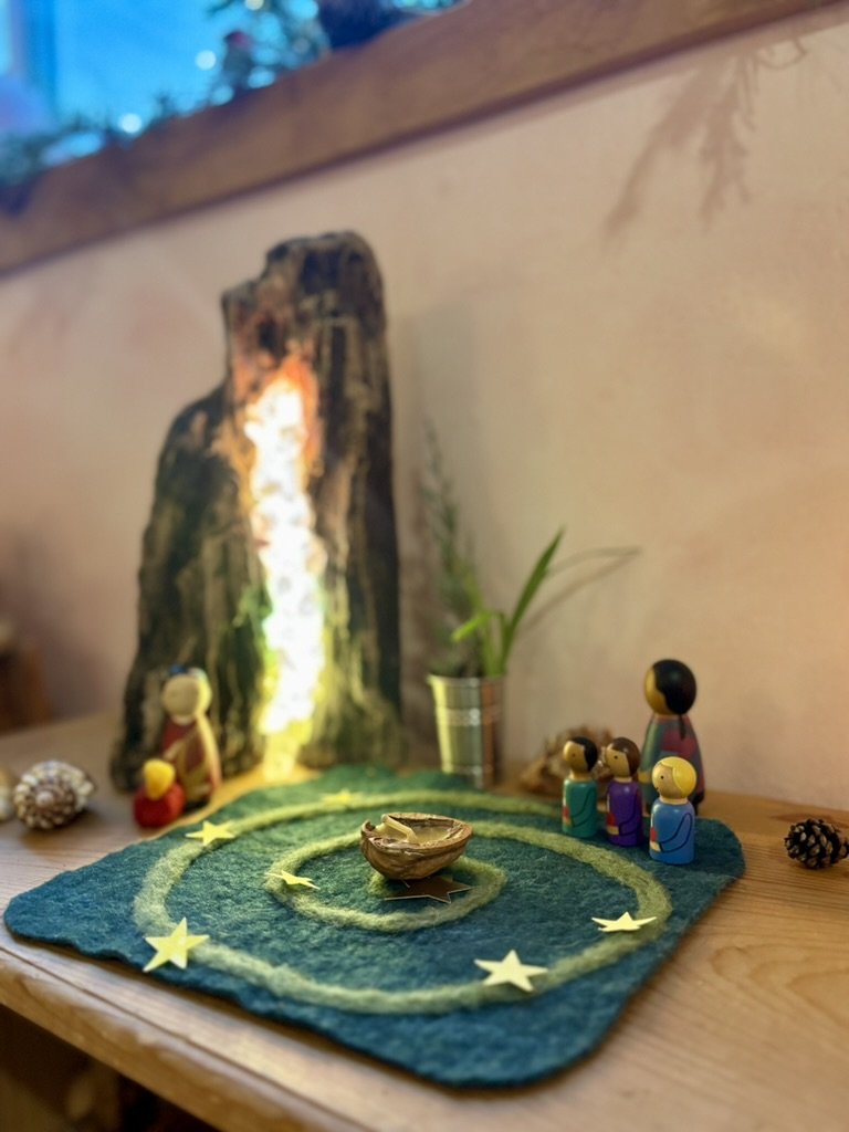 Nativity scene with wooden figures, a felt backdrop, star-shaped decorations, and a lighted mountain or tree in the background.