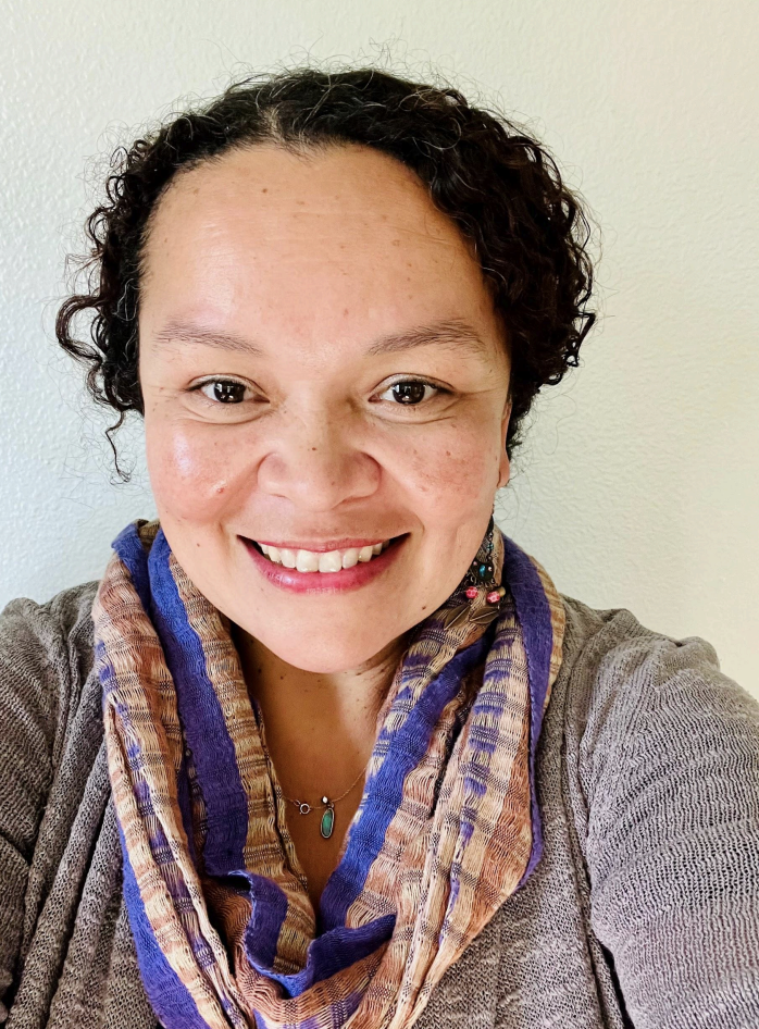 A woman with short curly hair smiling at the camera, wearing a multicolored scarf and a gray sweater, in front of a plain wall.
