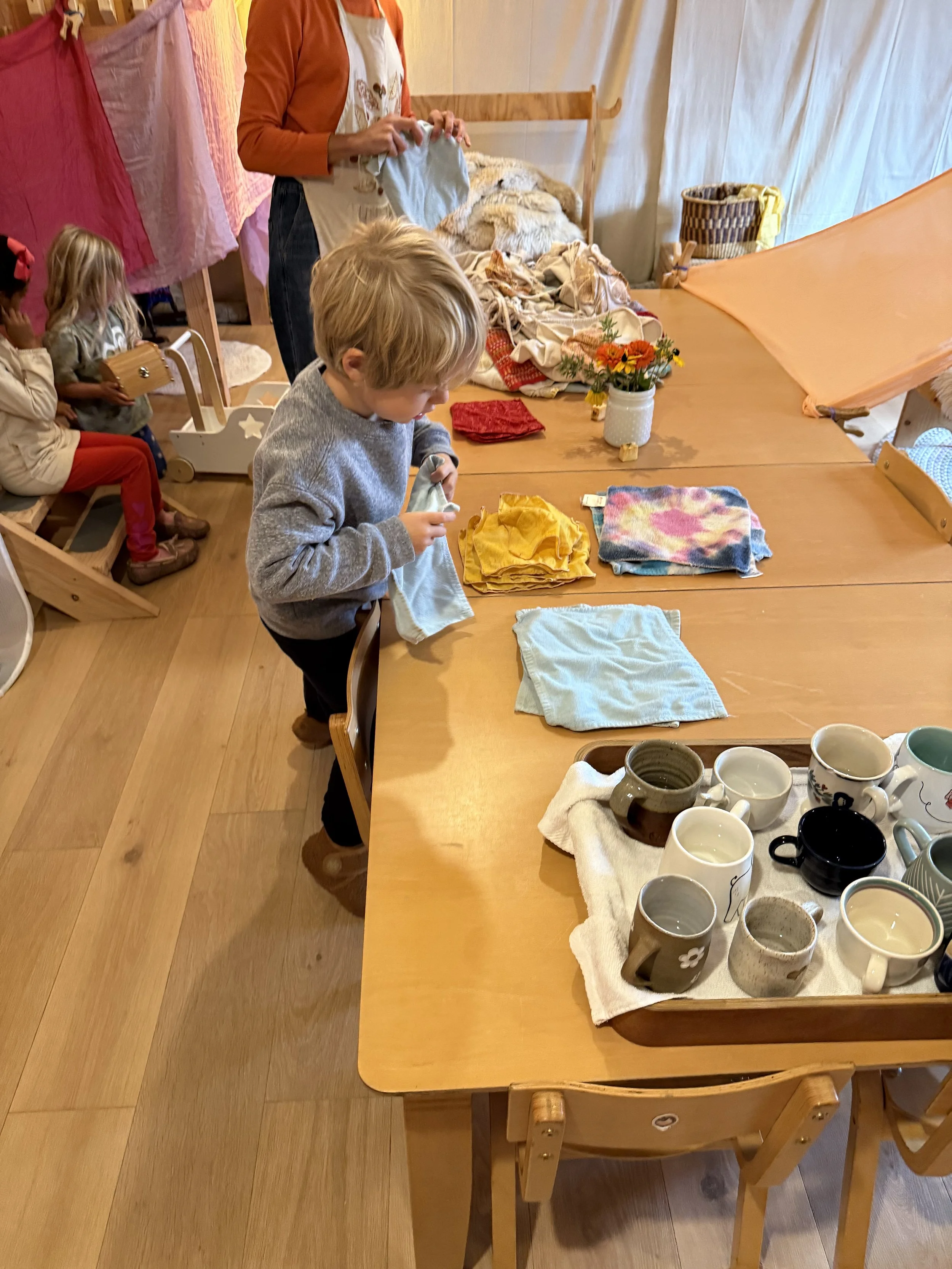 Children playing with laundry and dishes in a cozy, rustic room with wooden floors and a large table.
