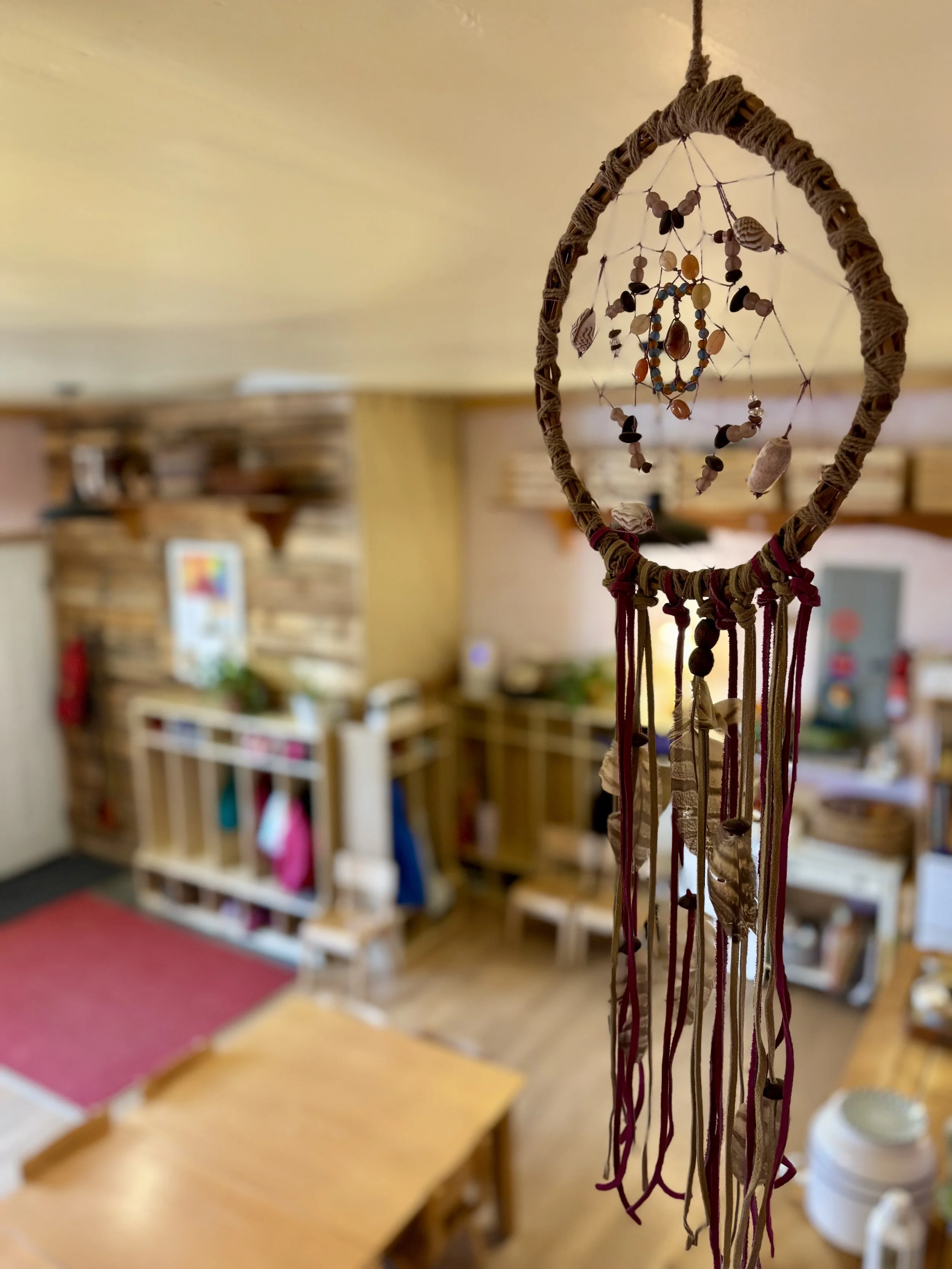 A dreamcatcher made with woven cord, beads, shells, and feathers hanging in a cozy home interior with wooden furniture and a colorful rug.