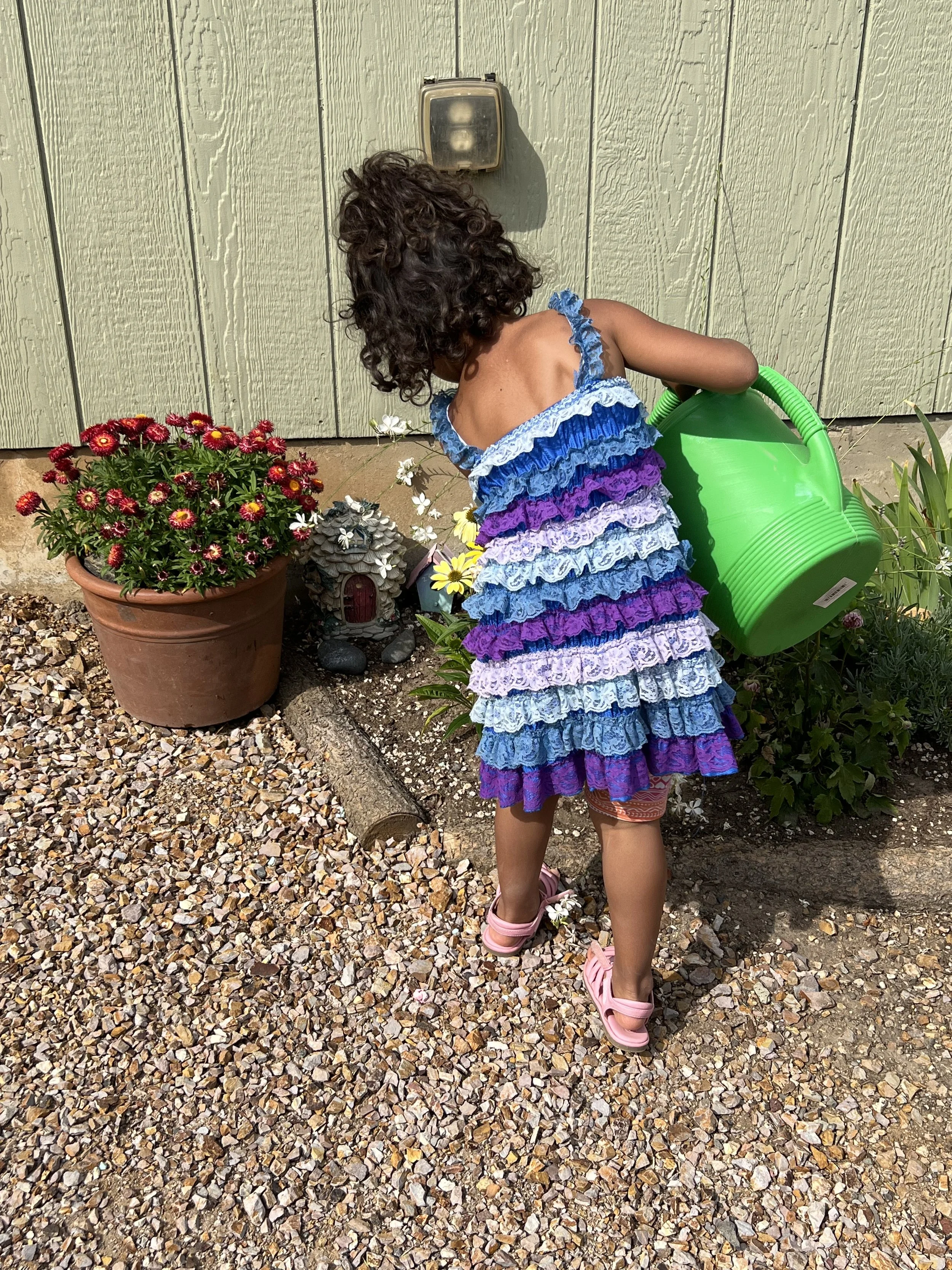 A young girl with curly hair wearing a colorful ruffled dress and pink sandals watering plants in a garden.