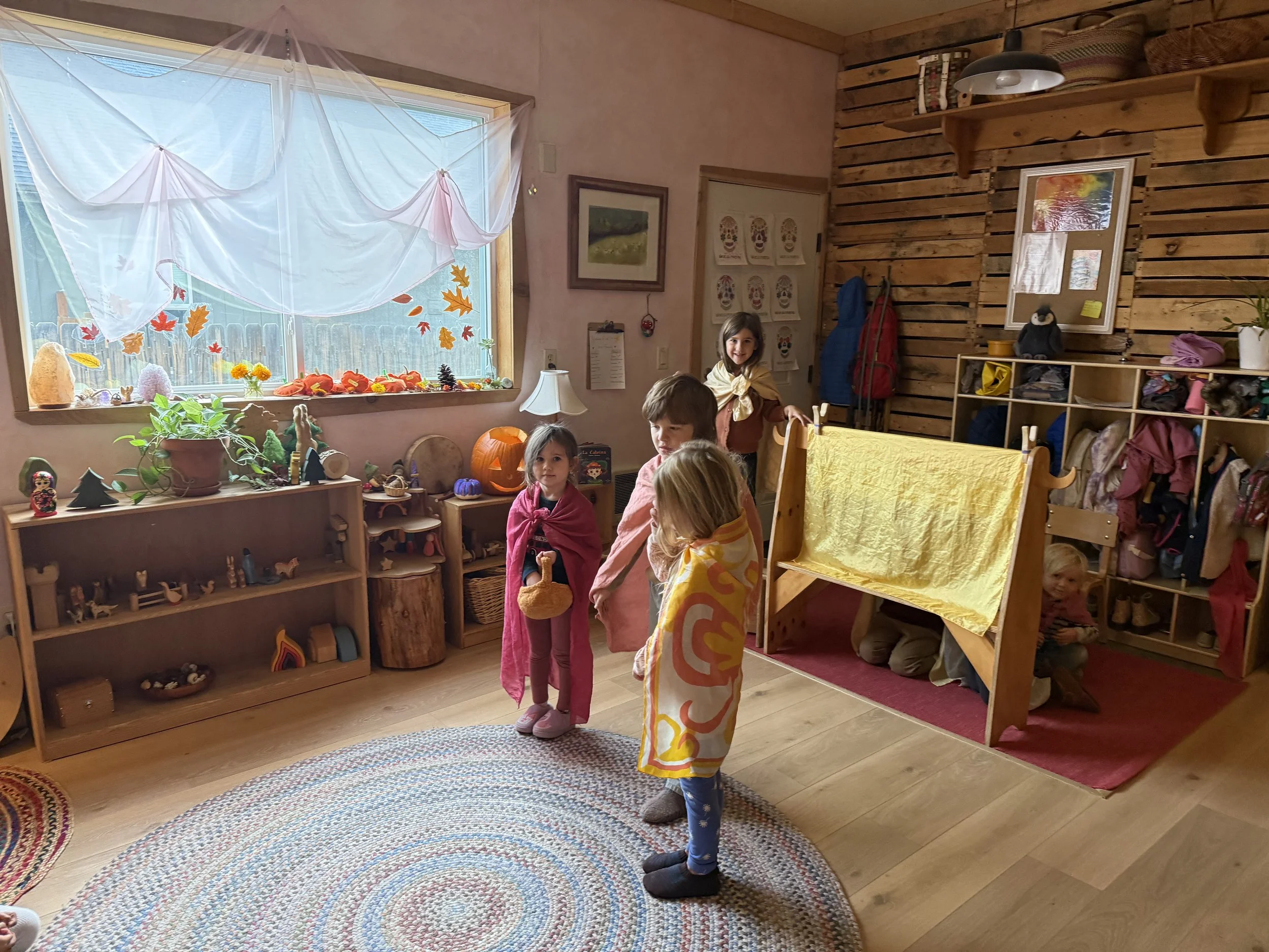 Children playing in a cozy indoor classroom decorated with autumn-themed crafts and decorations, with a large window and wooden shelves filled with toys and artwork.