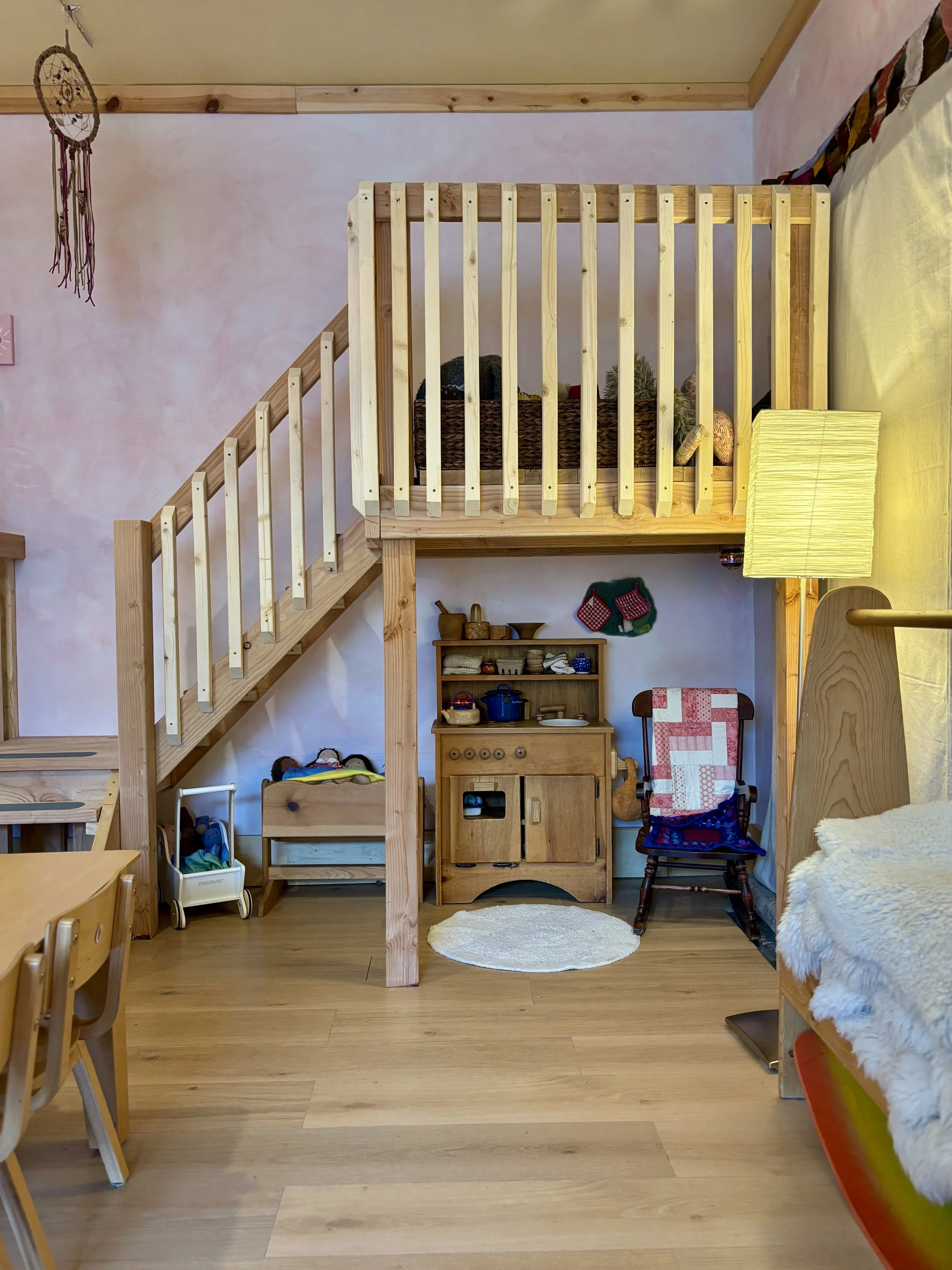 A cozy children's playroom with a wooden loft bed, stairs, and a small play kitchen under the loft. Soft lighting, a rocking chair with a blanket, a round rug, and various toys and decorations are visible.