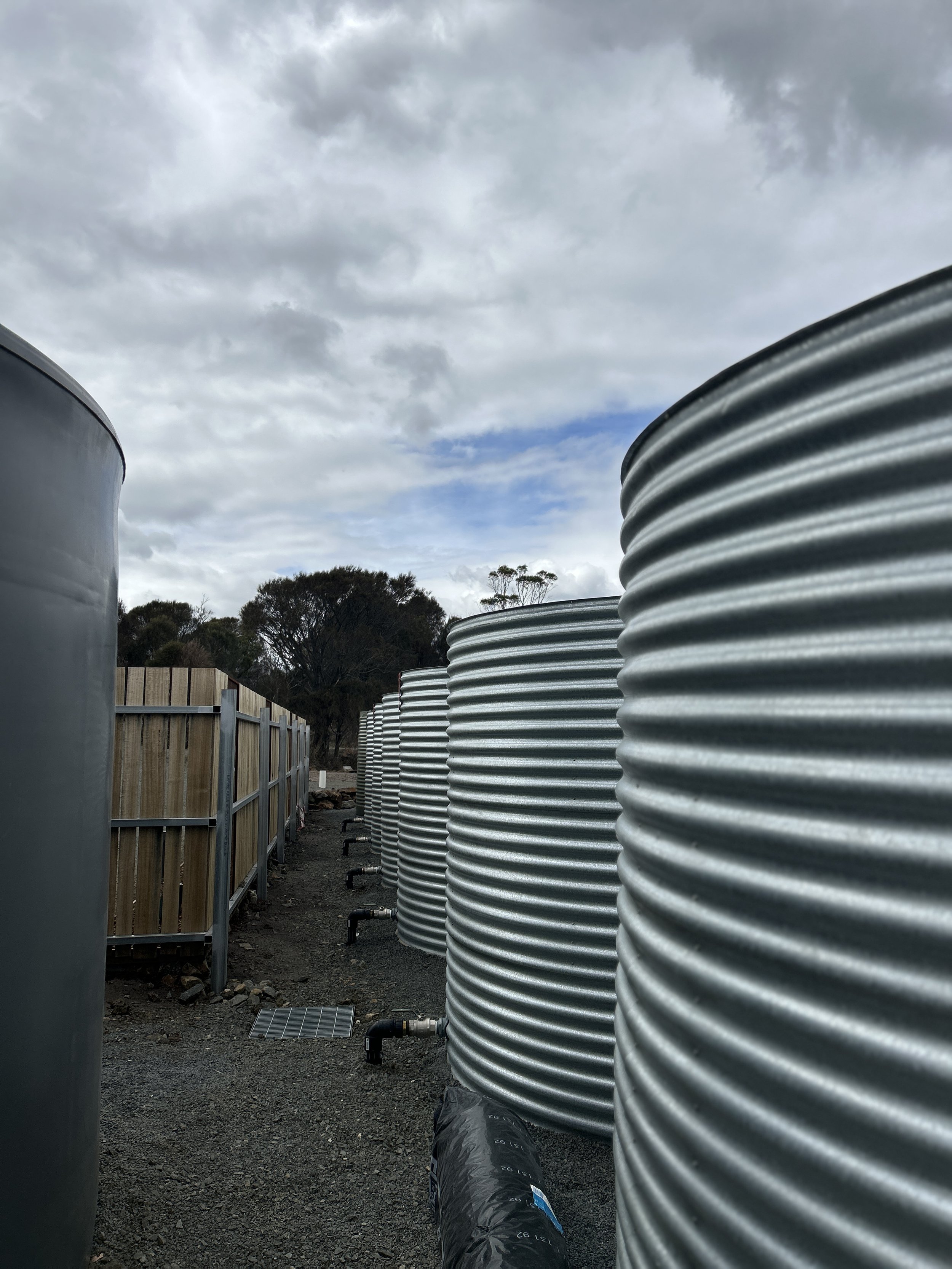 Row of large, corrugated metal water tanks outdoors under cloudy sky.