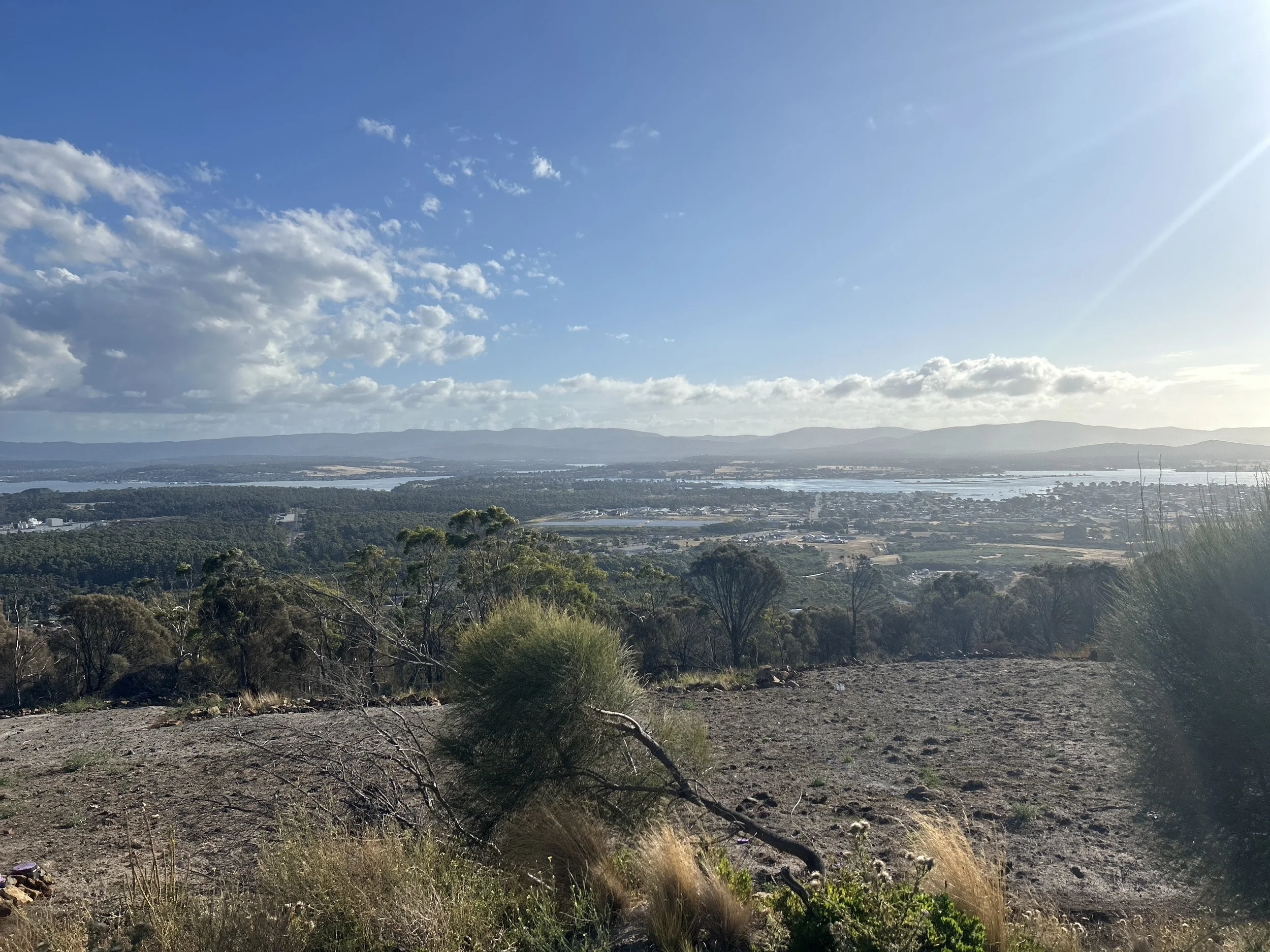 A scenic landscape view includes distant mountains, a large body of water, and a sky with scattered clouds. Foreground shows dry, rocky terrain with sparse vegetation and small bushes.