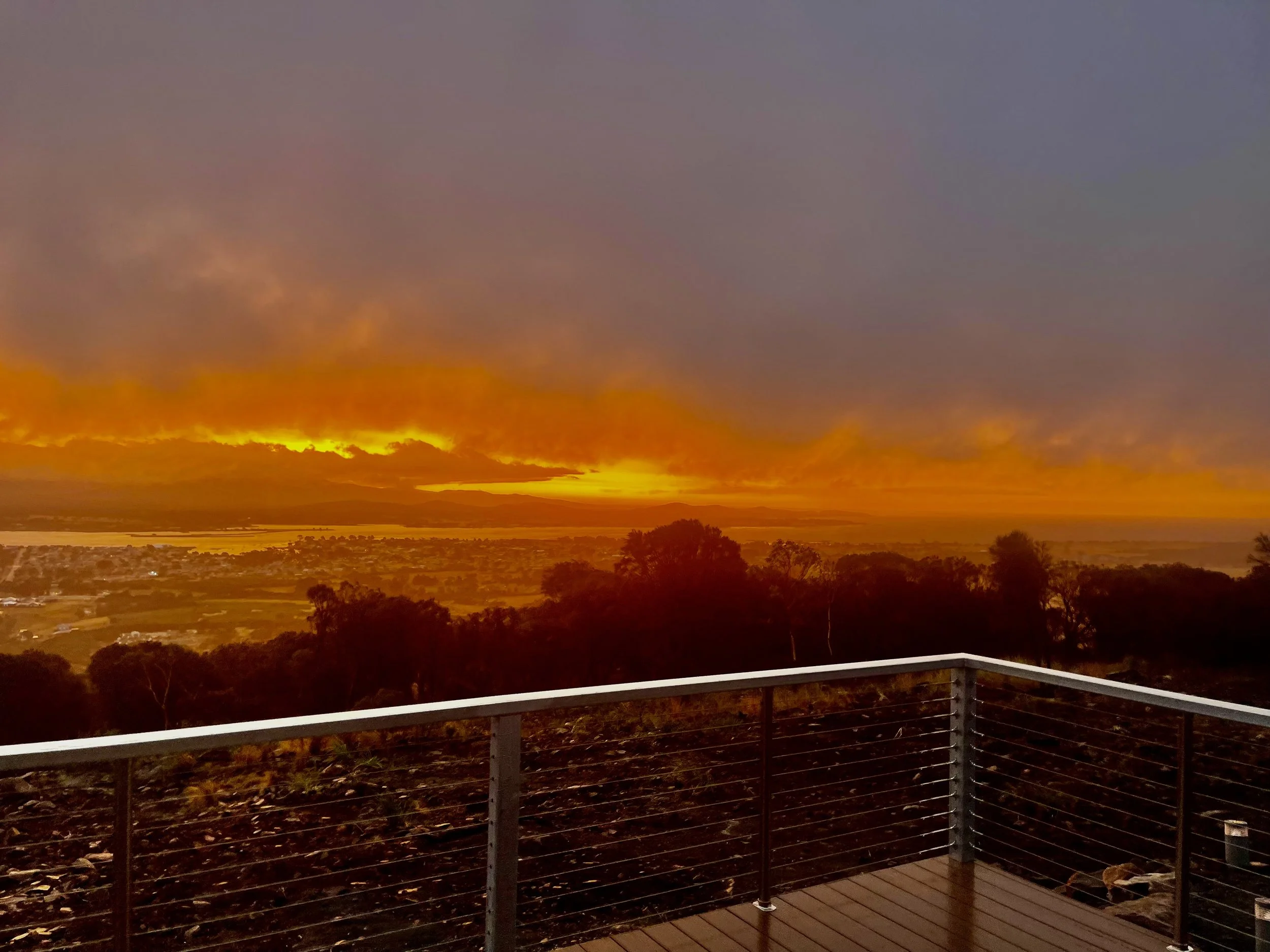 Sunset over a landscape with trees, water, and cloudy sky, viewed from a balcony with metal railing.
