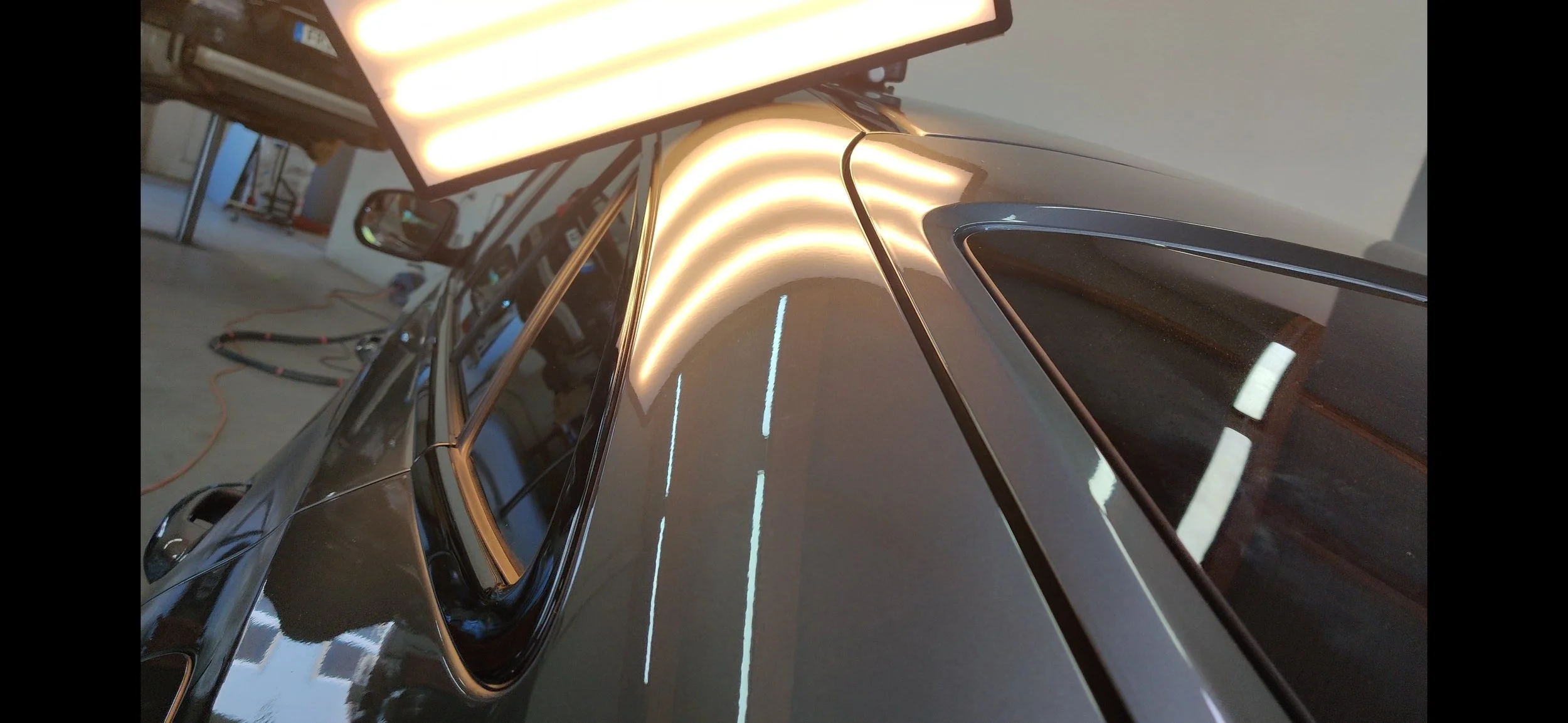 Nahaufnahme eines schwarzen Autos in einer Werkstatt, die Dachlinie und die Seitenscheiben sind sichtbar, die Umgebung hat Werkzeuge und Kabel im Hintergrund.