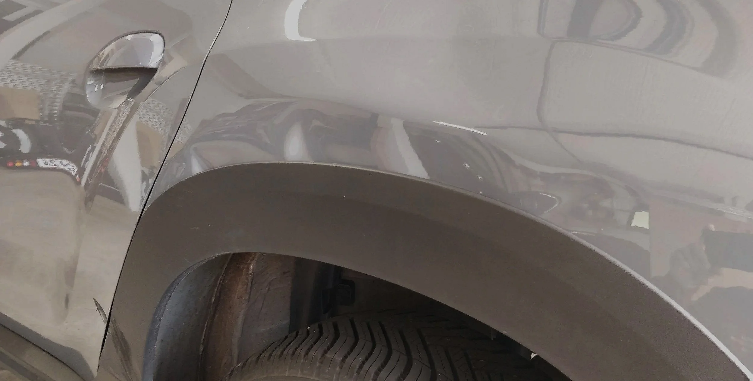 Nahaufnahme eines Fahrzeugkotflügels, Reifen, Felge und Fahrzeugseite, reflektierende Oberfläche, Parkhaus im Hintergrund.