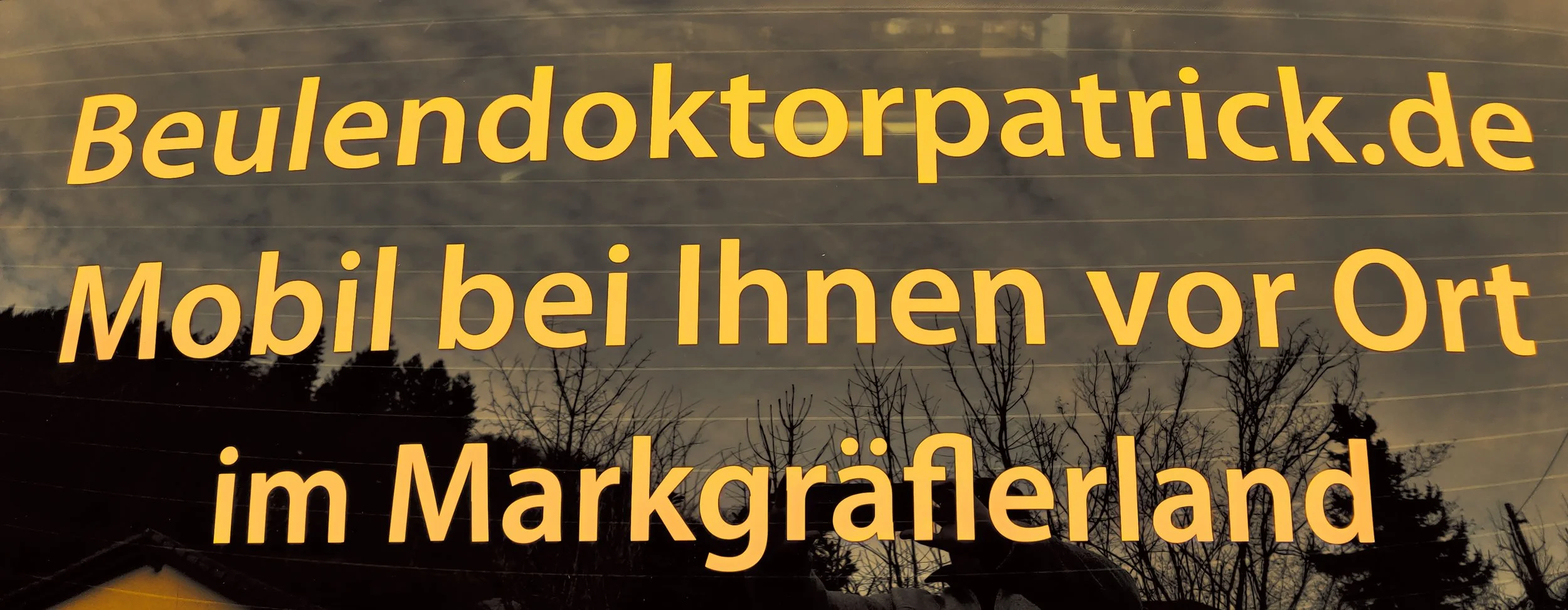 Schwarze Fensterbeschriftung mit gelben Buchstaben, die eine Werbebotschaft für einen mobilen Service vor Ort im Markgräflerland anzeigen, bei Dämmerung oder bewölktem Himmel