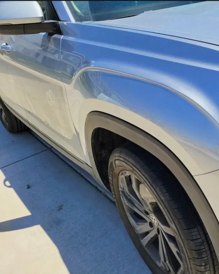 Nahaufnahme eines blauen Autos mit leichter Delle an der Beifahrertür, parkend auf einem glatt betonartigen Boden.