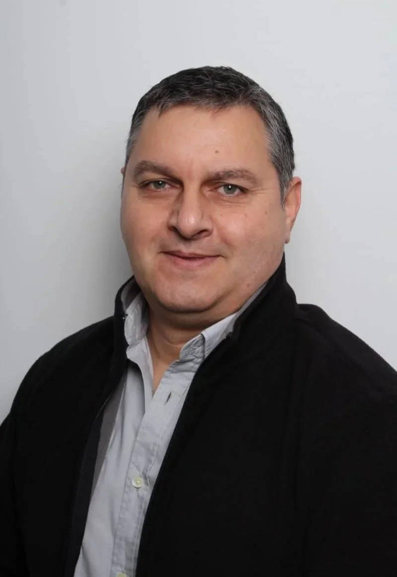 Porträt eines Mannes mit dunklem Haar, blauer Augen, trägt einen schwarzen Pullover über einem weißen Hemd, vor einem weißen Hintergrund.