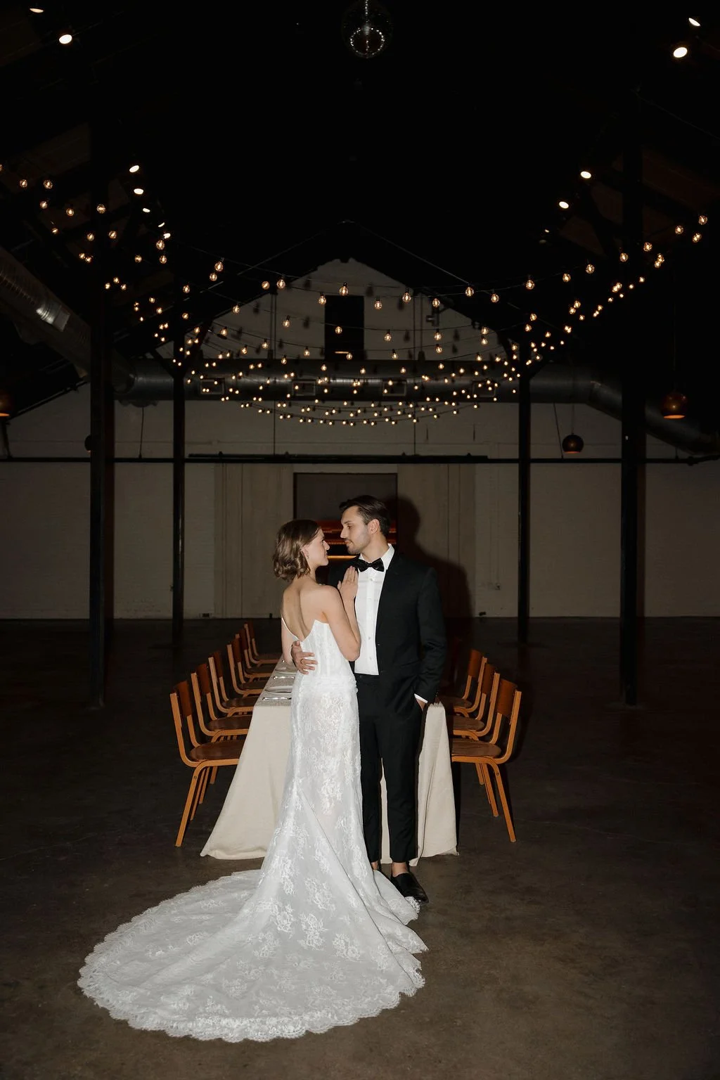 A bride and groom standing in front of a reception table and string lights hung above them.