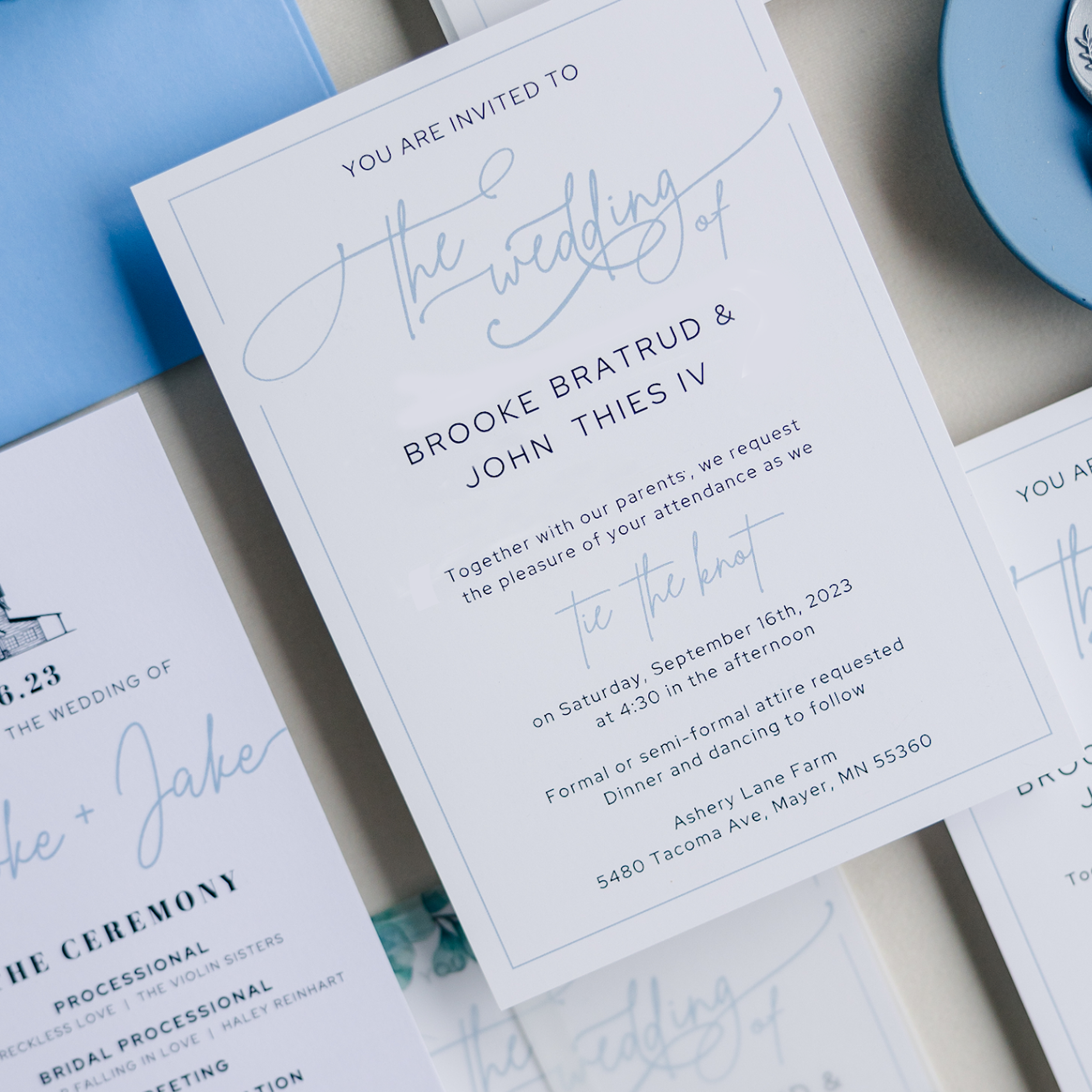 A white wedding invitation with blue text laying on top of a blue envelope and a ceremony program.