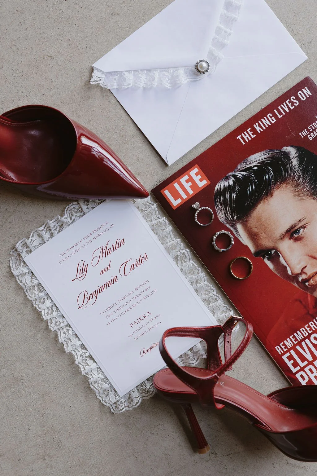 White wedding invitation with lace trim surrounded by a red magazine, wedding rings, and red heels