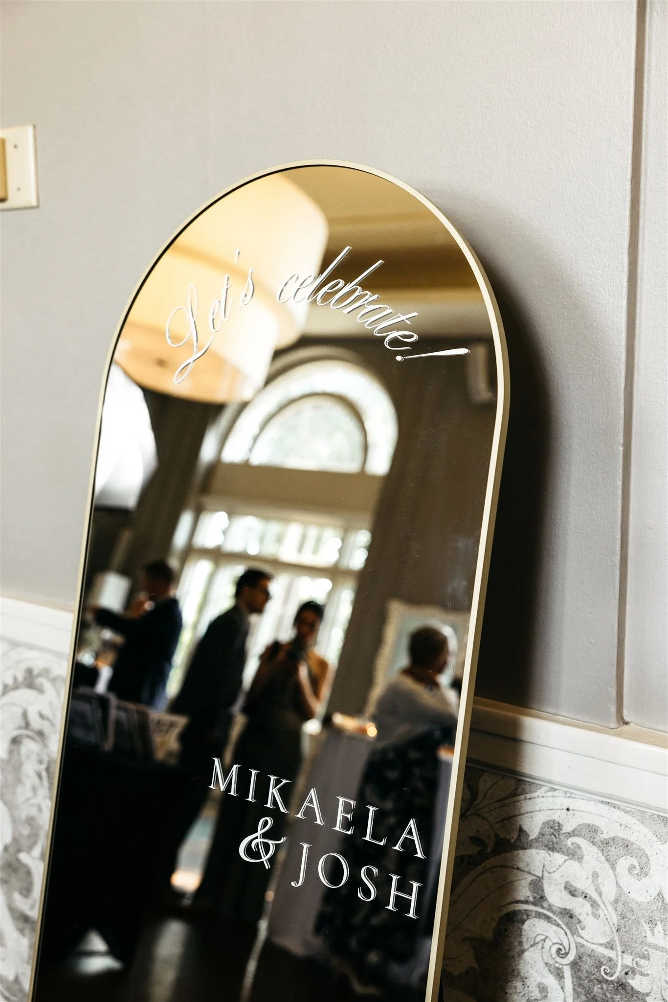 A mirror in an elegant wedding venue with the words "Let's celebrate!" and "Mikaela & Josh" in white text.