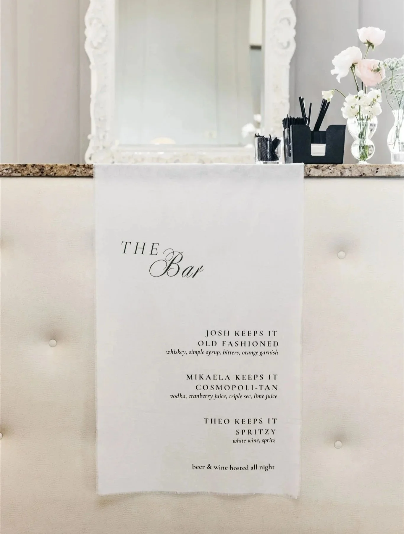Menu titled 'The Bar' on a white cloth with drink options and ingredients, set against a white background with a mirror and flowers.