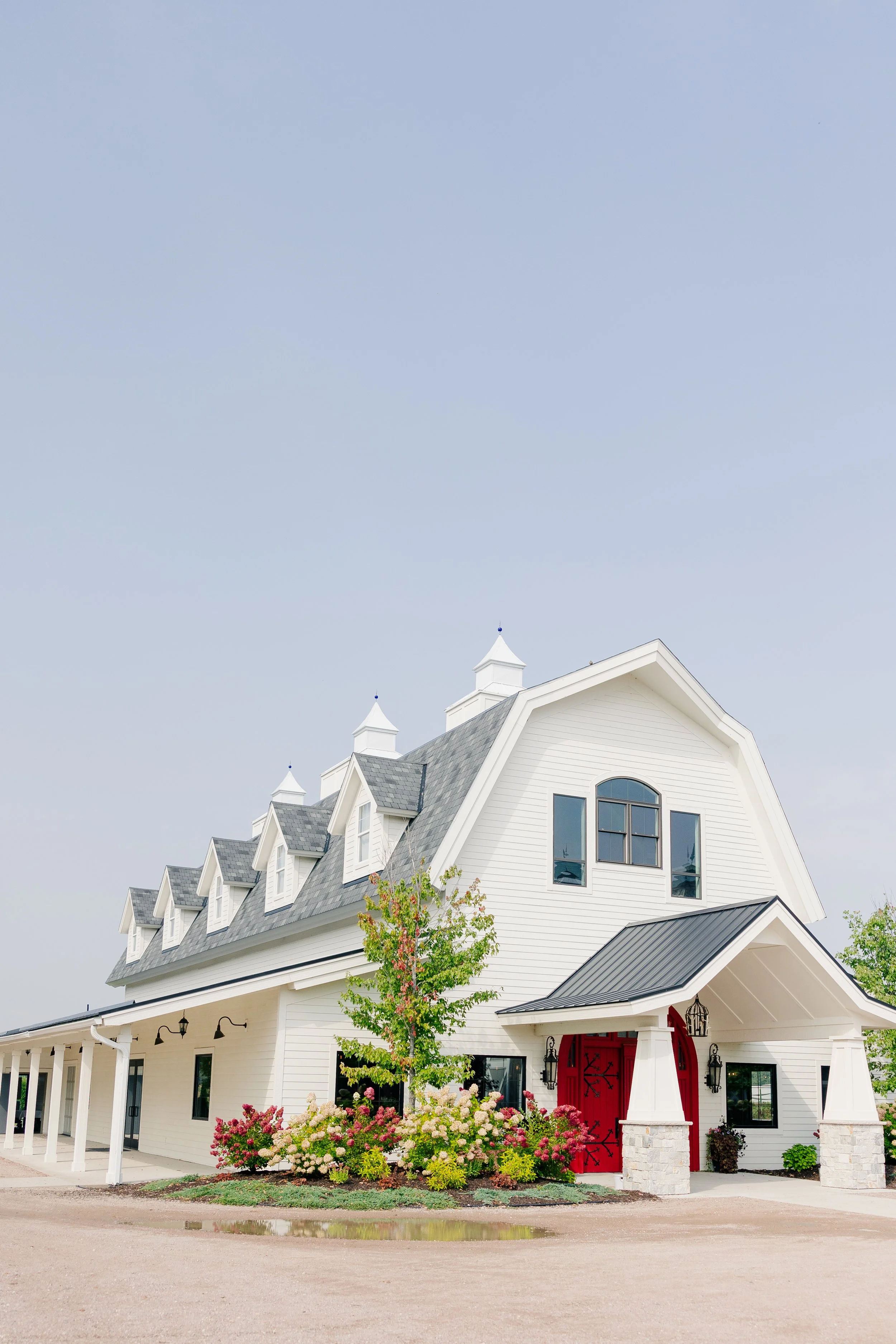 A large, white, three-story barn-style house with a gray roof and a red front door, surrounded by colorful bushes and a small tree against a blue sky.