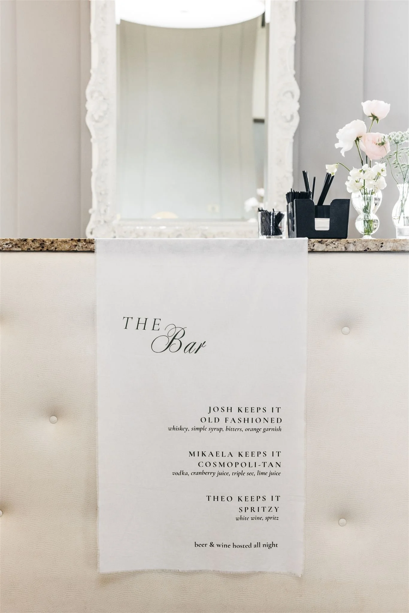A menu titled 'The Bar' hanging on a white cushioned wall, listing drinks and their ingredients, with a countertop holding black straws, stirrers, and floral arrangements in vases.