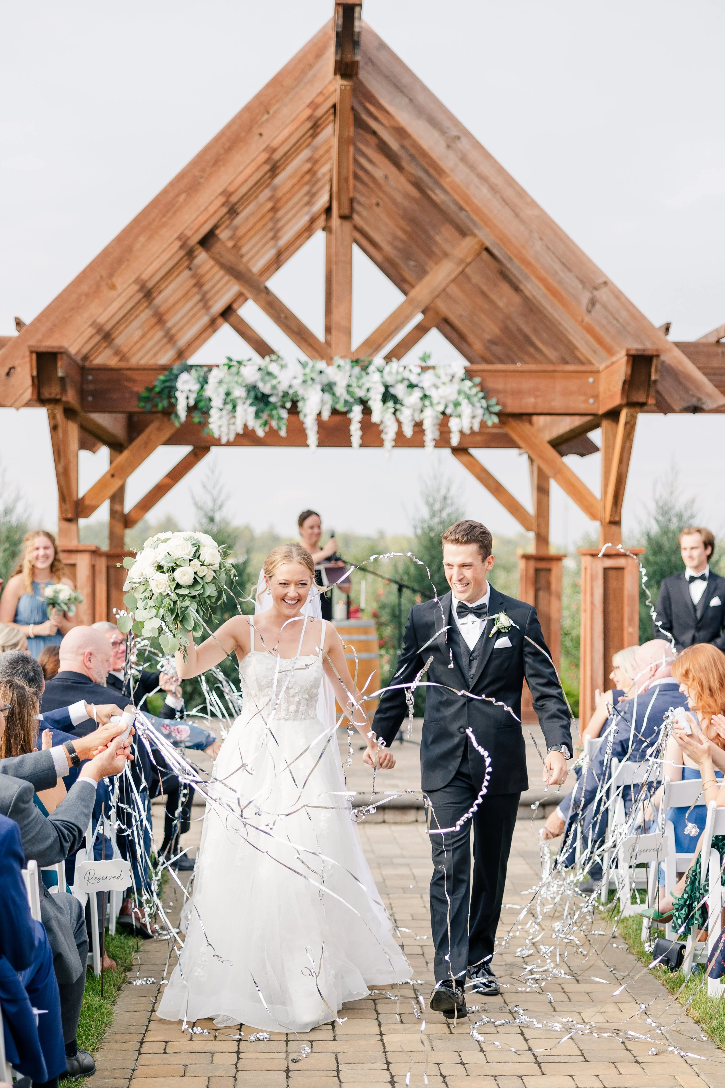 Bride and groom walking down the aisle at a wedding ceremony, smiling with guests seated on both sides, under a wooden arch decorated with white flowers.
