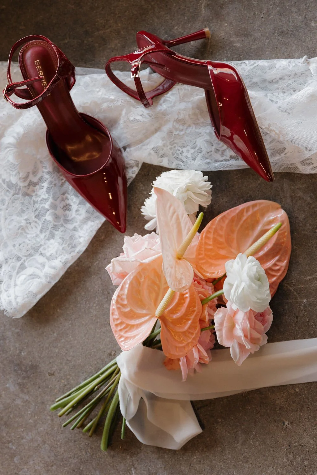 A bouquet of pink flowers laid on a concrete floor next to red heels and a lace wedding dress.