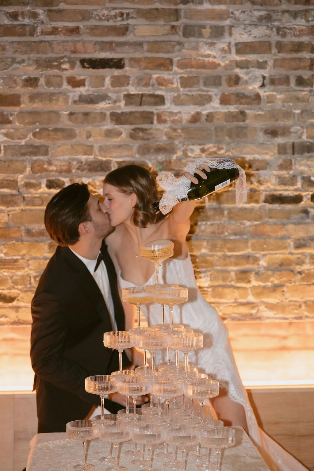 A bride and groom pouring champagne onto a champagne glass tower while kissing with a brick wall behind them.