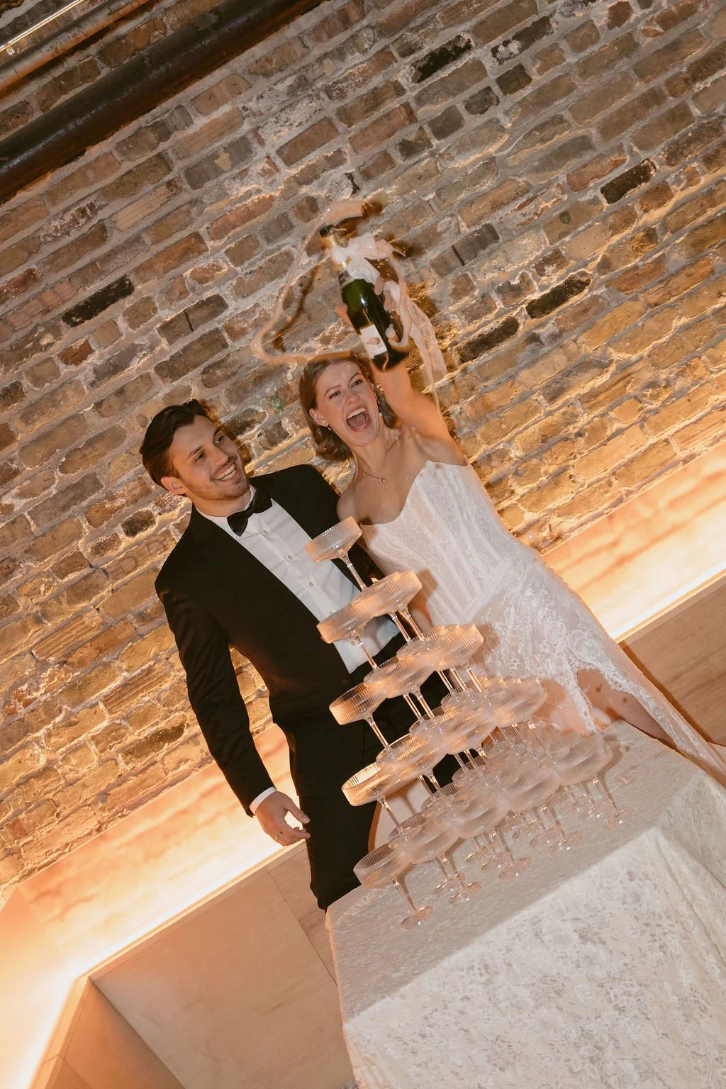A bride and groom popping a champagne bottle by a champagne glass tower while smiling with a brick wall behind them.