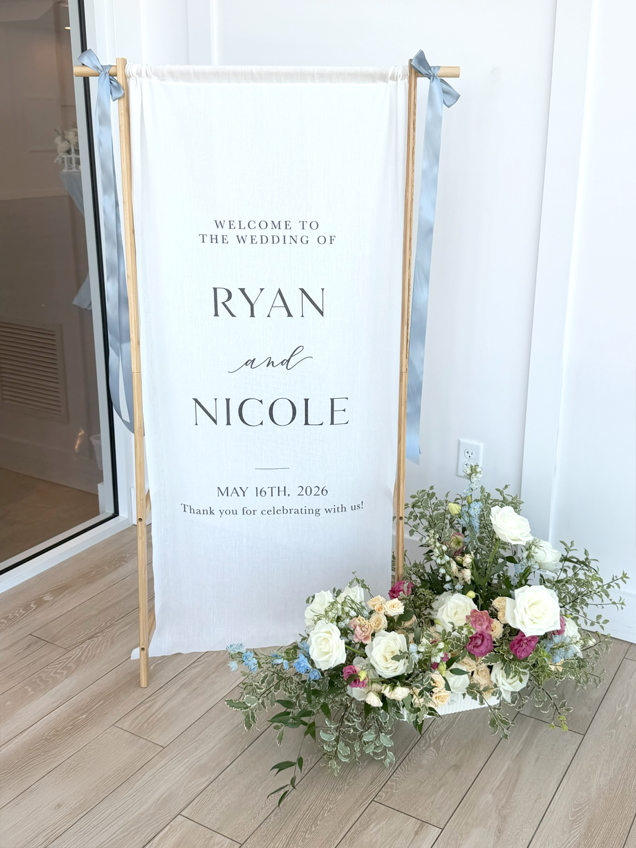 White fabric sign with blue ribbon hanging from a wooden stand and a flower arrangement in the front.