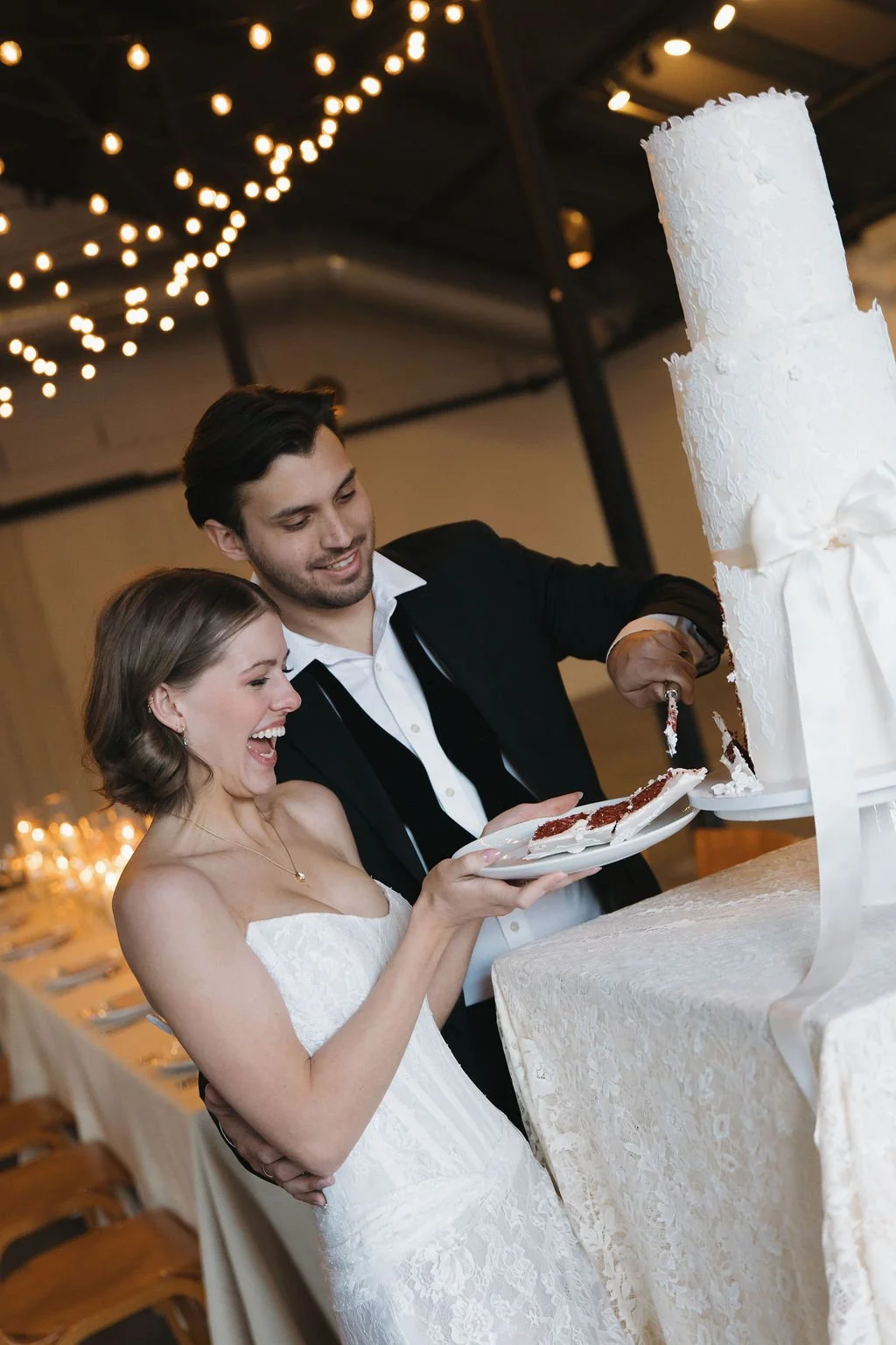 A bride and groom cutting a wedding cake with a reception table and string lights behind them.