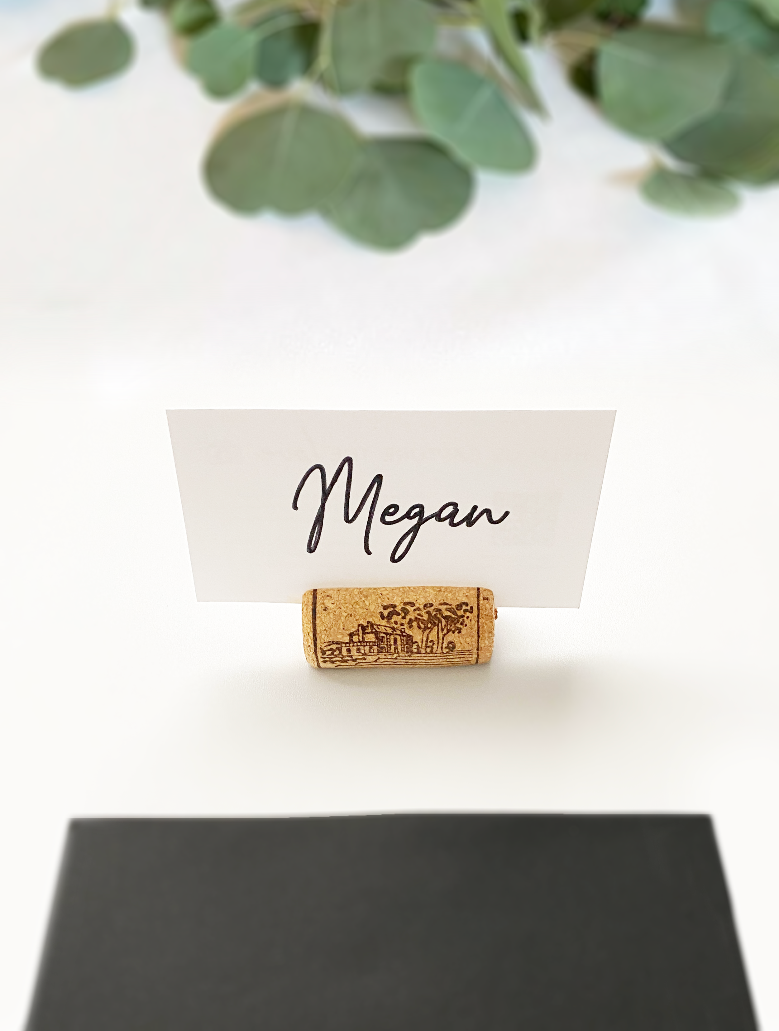 A printed place card with the name "Megan" in cursive text sitting in a wine cork holder on a white tablecloth with eucalyptus branches in the background.