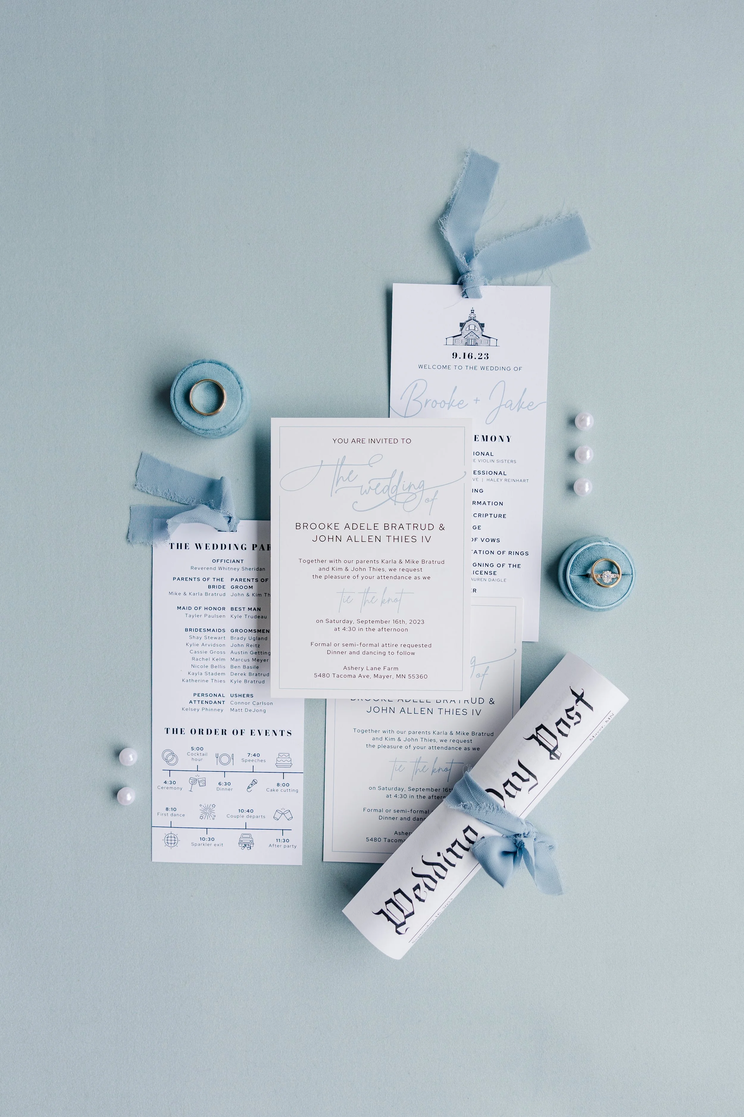 Wedding invitation suite including an invitation card, ceremony program, and a wedding newspaper scroll tied with blue ribbon, along with a wedding ring and small decorative pearls on a light blue background.