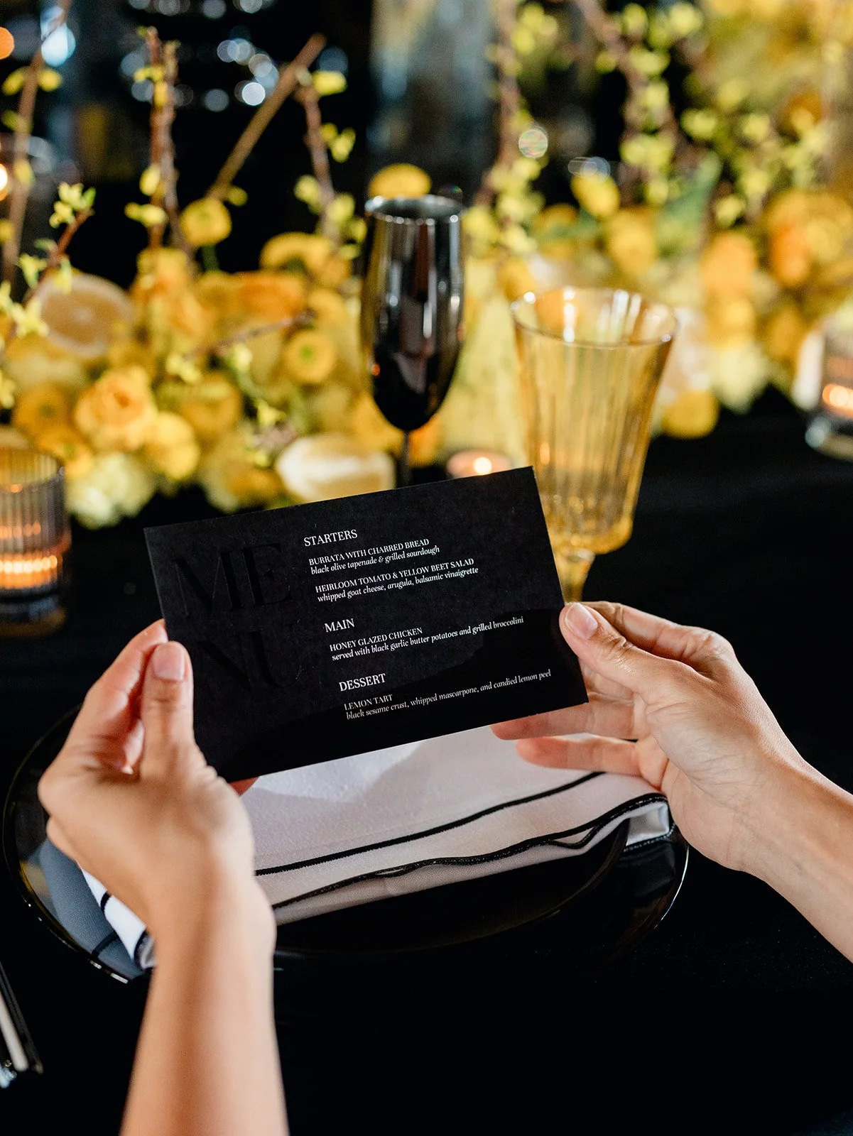 A black and white dinner menu being held over a place setting with a full display of yellow flowers in the backgorund.