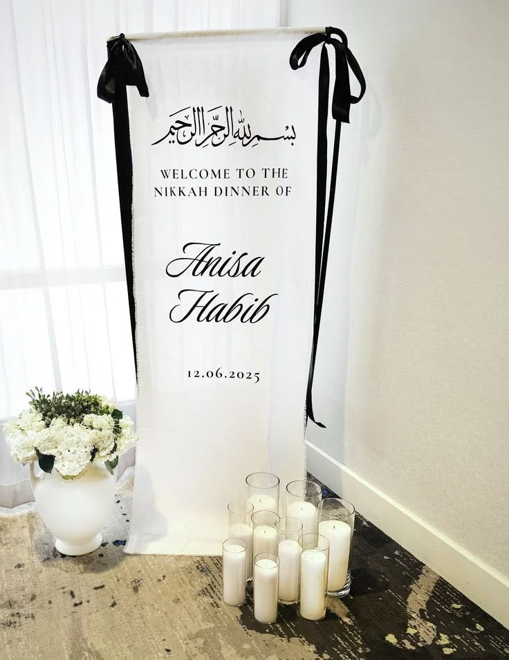 A white fabric welcome sign with black ribbon and the message "Welcome to the Nikkah Dinner of Anisa Habib" on it