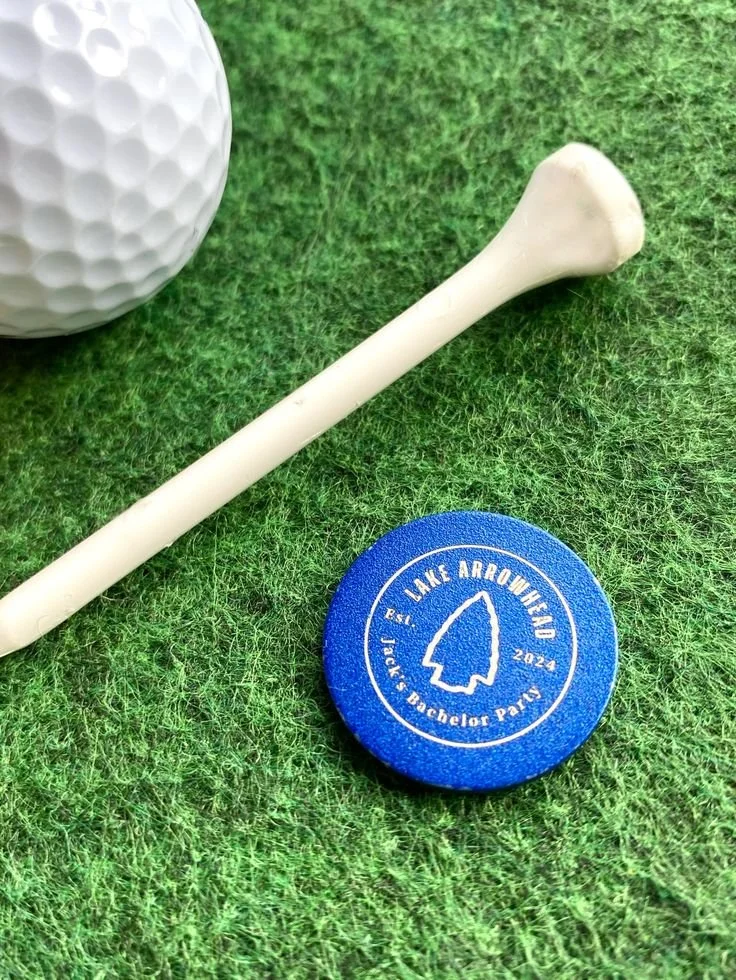 Blue ball marker with a custom design laying on the grass next to a golf ball and tee.