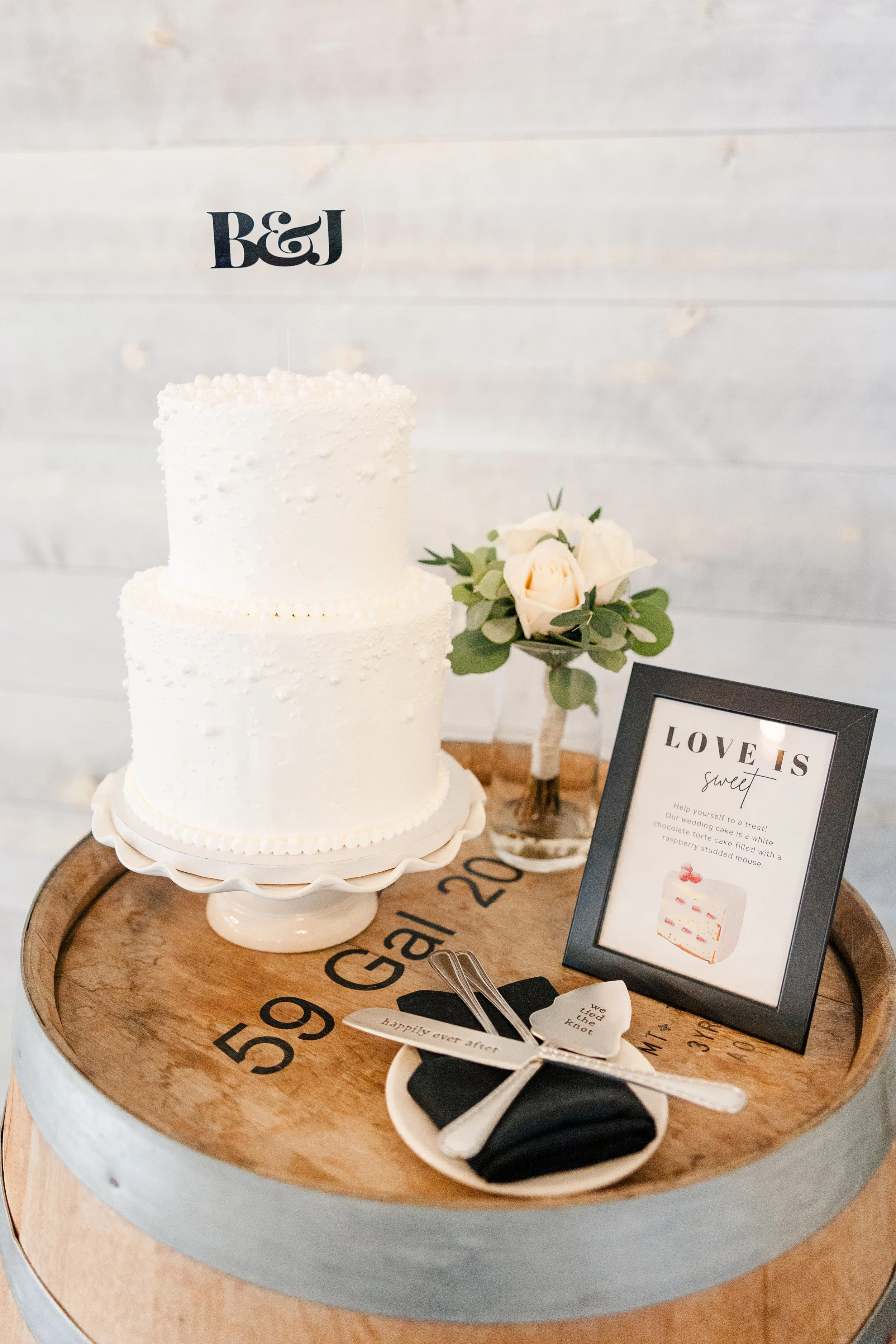 A two-tiered white wedding cake on a cake stand, decorated with small white dots. Next to the cake is a small bouquet of white roses with greenery, a framed sign that reads 'Love is sweet' with an image of a cake, and a plate with wedding utensils wr